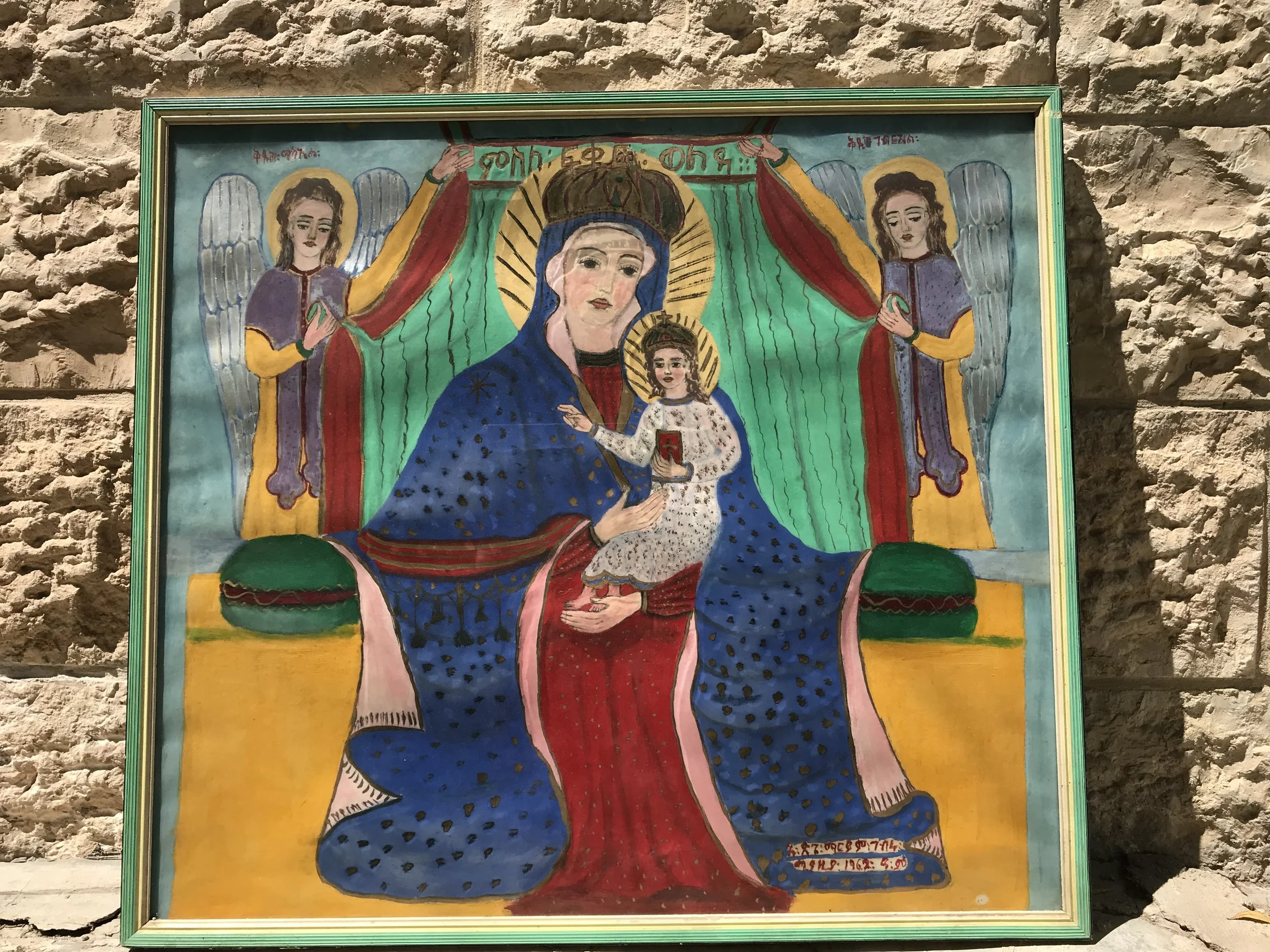 A colorful religious painting of the Virgin Mary with baby Jesus, flanked by two angels, set against a bright yellow background.