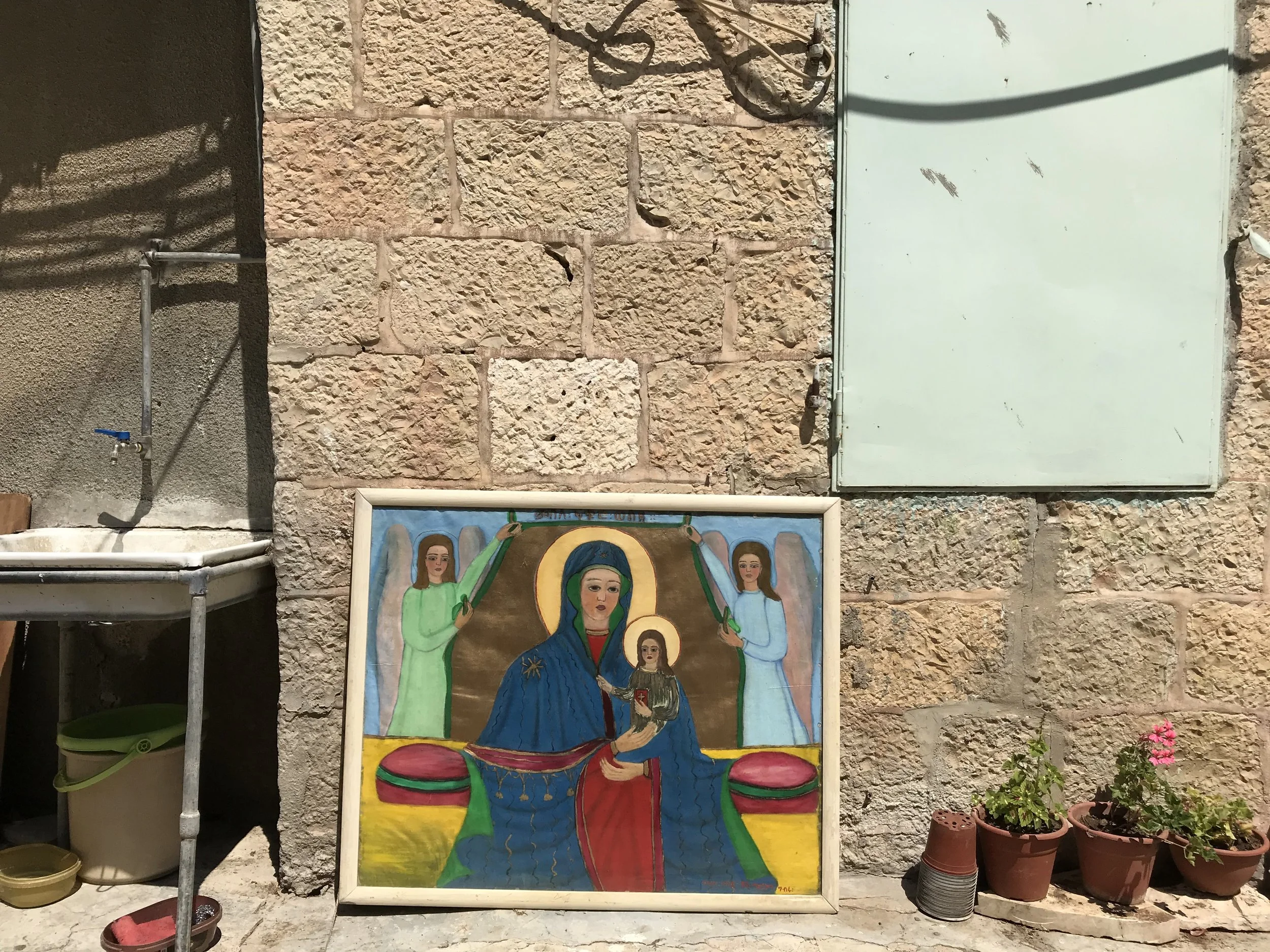 Colorful religious icon painting of the Virgin Mary holding baby Jesus, flanked by two angels, placed against a stone wall with plants and pots nearby.
