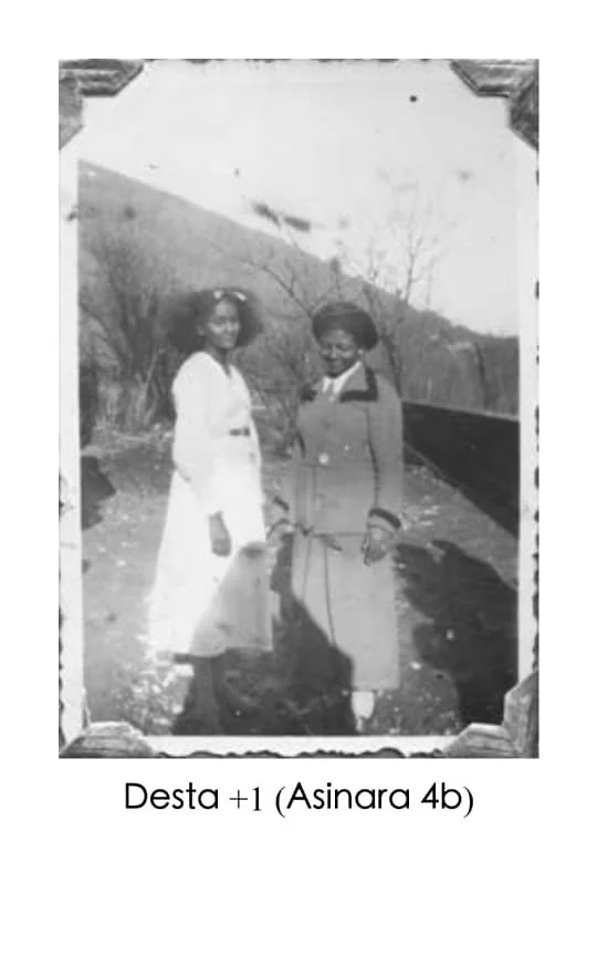 Two women standing outside on a sunny day, with trees and hills in the background, dressed in vintage clothing from the mid-20th century.