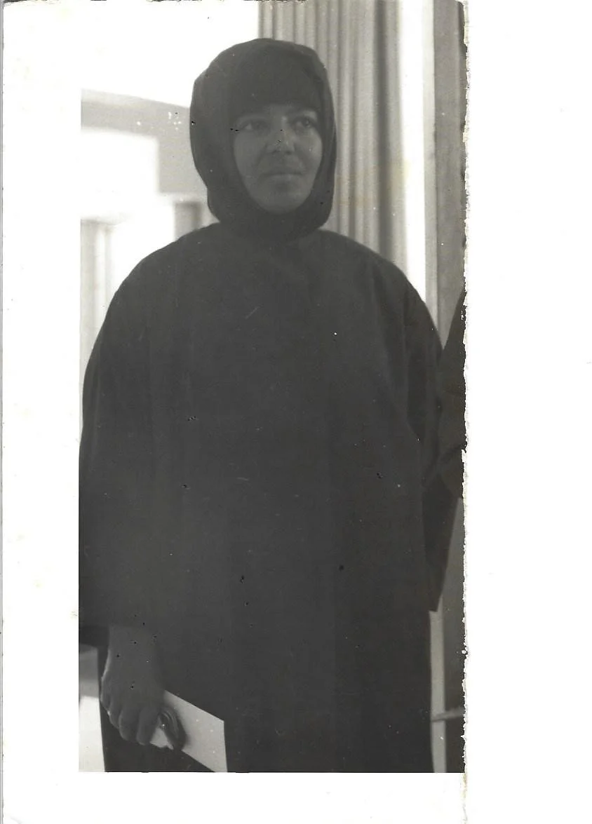 A woman wearing a hijab and dark clothing, standing indoors and holding a piece of paper or notebook in her right hand.