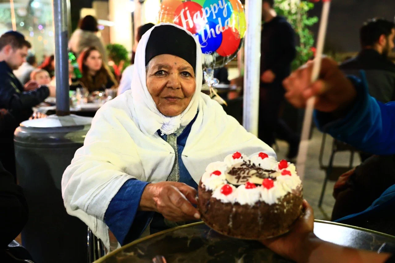 An elderly woman in a white headscarf sitting at a table during a birthday celebration, being presented with a chocolate cake decorated with whipped cream, cherries, and chocolate pieces, at night in a well-lit indoor or outdoor space with people in 