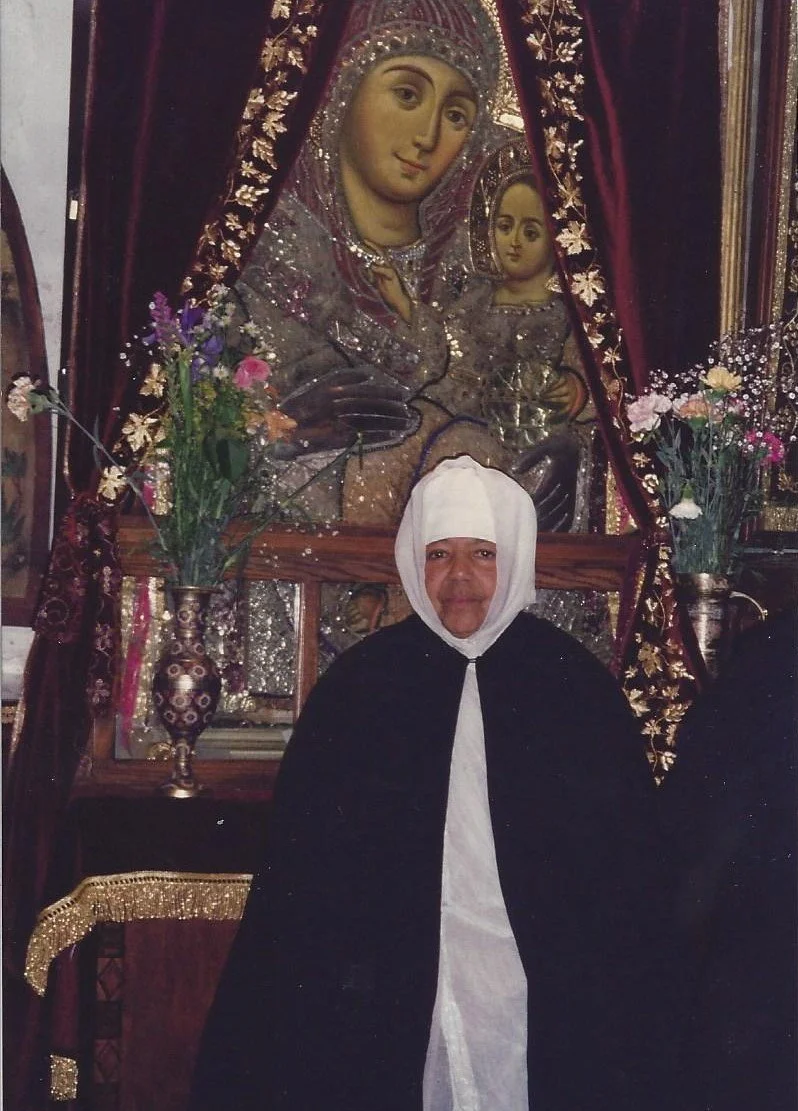 A woman wearing a white head covering and black garment standing in front of a large religious icon of the Virgin Mary and Child Jesus, decorated with flowers and drapes.