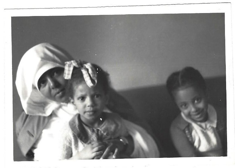 Black and white photo of a woman wearing a headscarf and glasses, sitting with two young girls smiling at the camera.