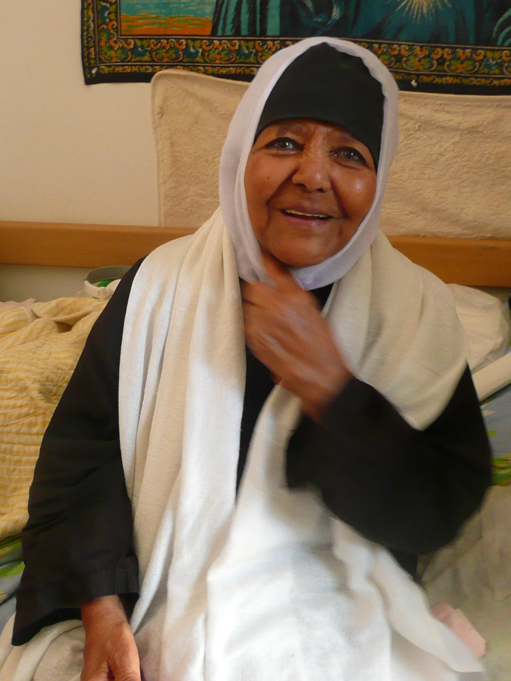 A smiling elderly woman wearing a black headscarf and a white shawl, sitting in a room with a colorful tapestry hanging behind her.