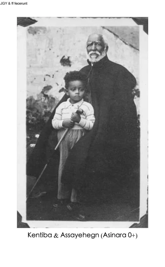 An old black and white photograph of an elderly man with a beard and glasses, sitting next to a young boy holding a stick. The background shows a wall with some damage. The caption reads: 'Kentiba & Assayehegn (Asina 0+)'.
