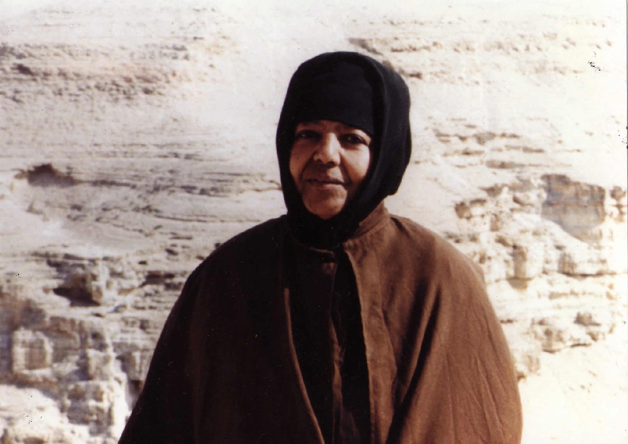 A person dressed in winter clothing, including a black hat and brown coat, standing outdoors in front of a snow-covered landscape.