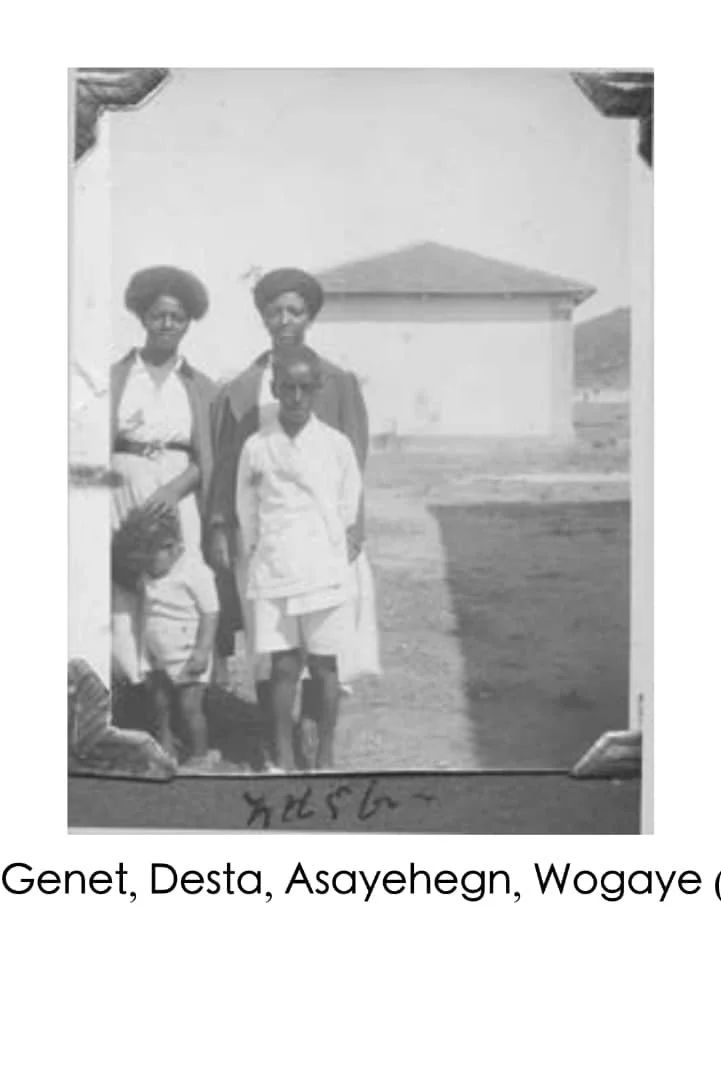 Black-and-white photo of four people standing outdoors in front of a building, with a handwritten message or signature at the bottom.