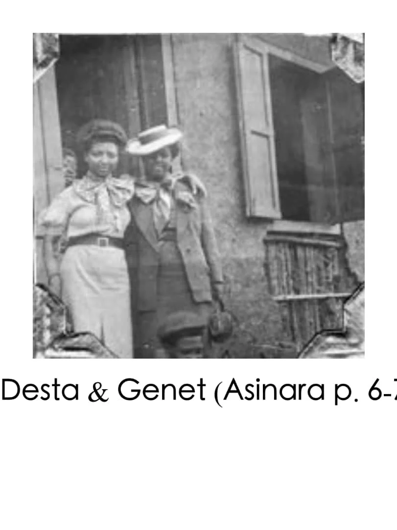Black and white photograph of a woman and man standing outside a building with open window shutters. The woman is dressed in a blouse and skirt with her arm around the man's shoulder. The man is wearing a hat, coat, and tie.
