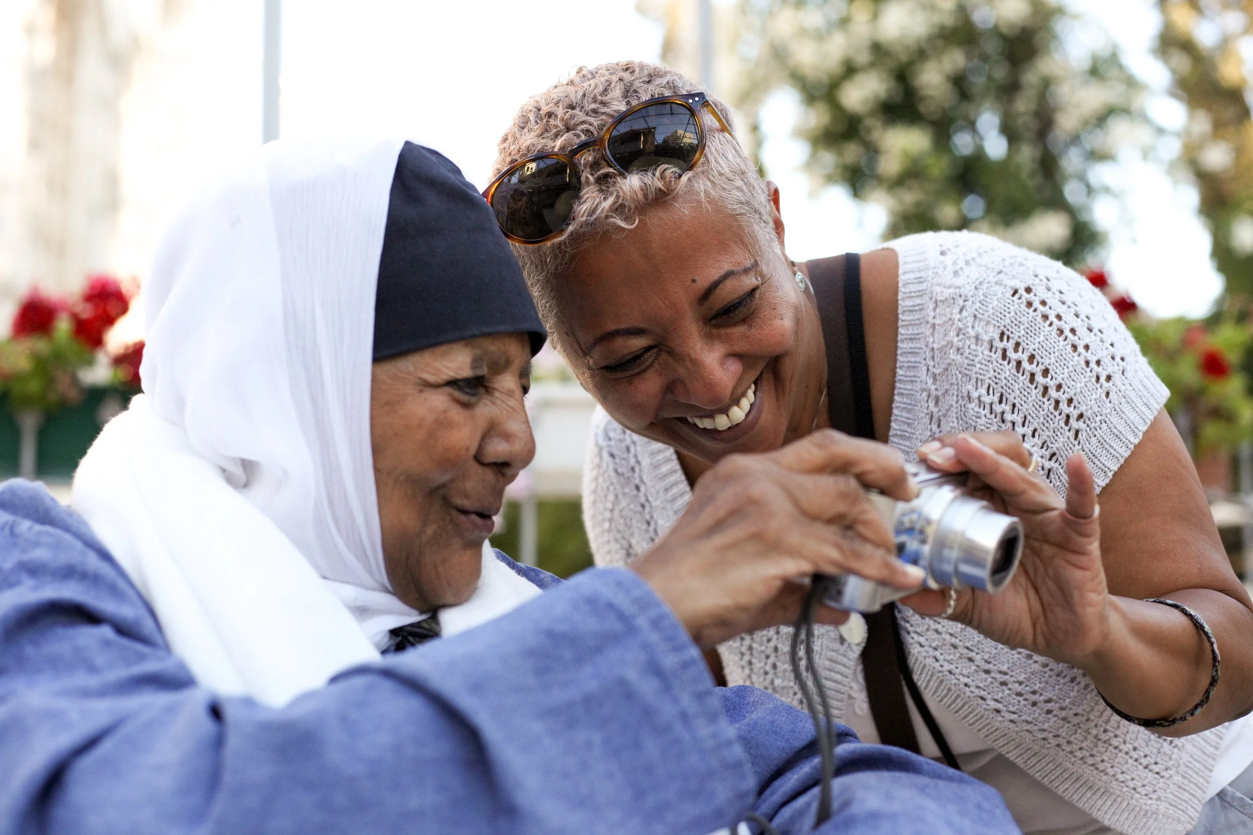 Two women smiling and looking at a camera outdoors, one is wearing sunglasses on her head, and the other is wearing a hijab.