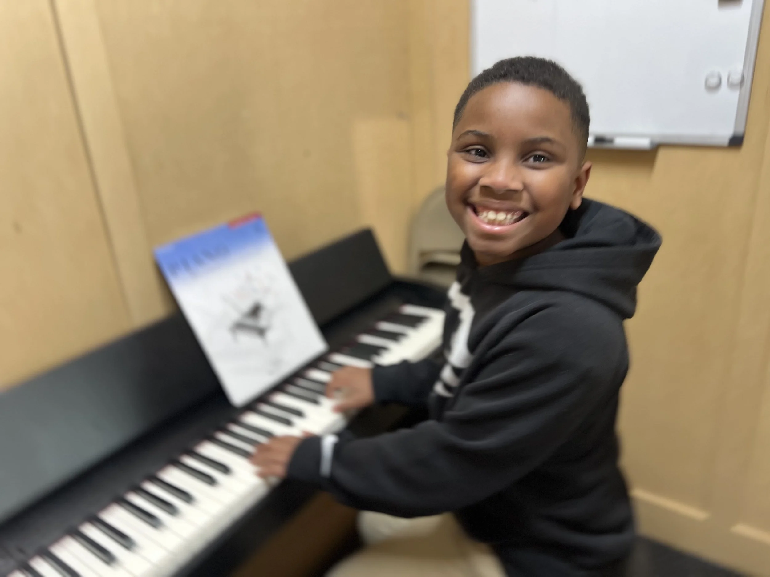 A smiling young boy playing a black and white piano in a classroom.