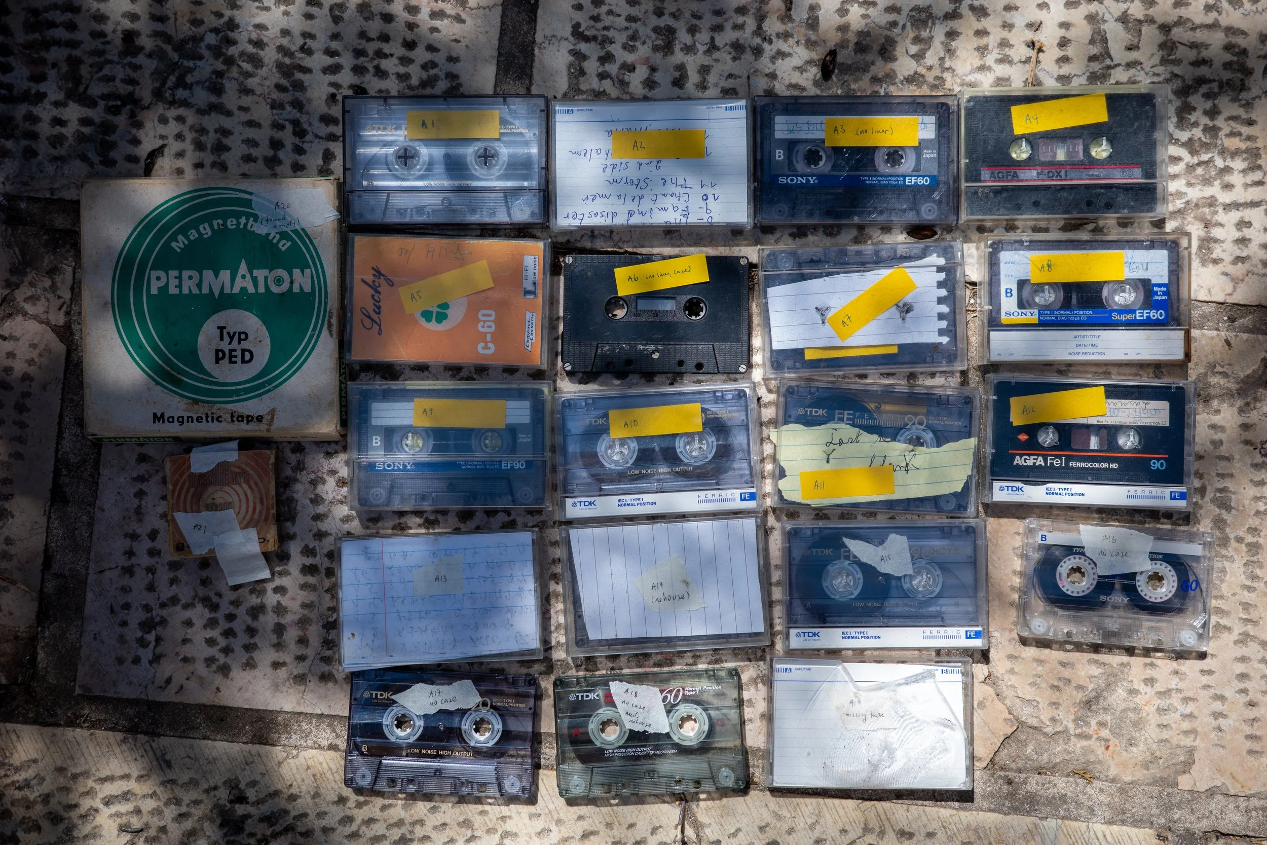 Multiple cassette tapes and a magnetic tape box are laid out on a granite surface. The tapes have handwritten labels with various notes and yellow tape labels with codes like A1, A2, A3, A4, A6, A7, A8, A9, A10, and A13. There are also lined paper no