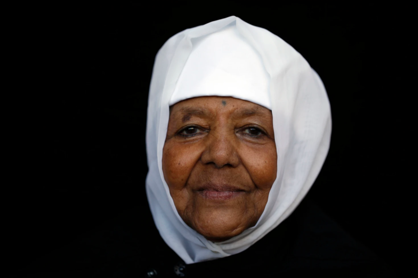 Close-up portrait of a smiling elderly woman wearing a white headscarf against a black background.