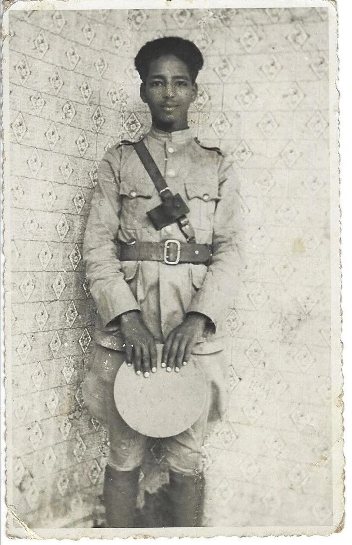 A young person in a military-style uniform with a hat, standing against a patterned wall, holding a round hat in one hand and carrying a camera strap across their chest.