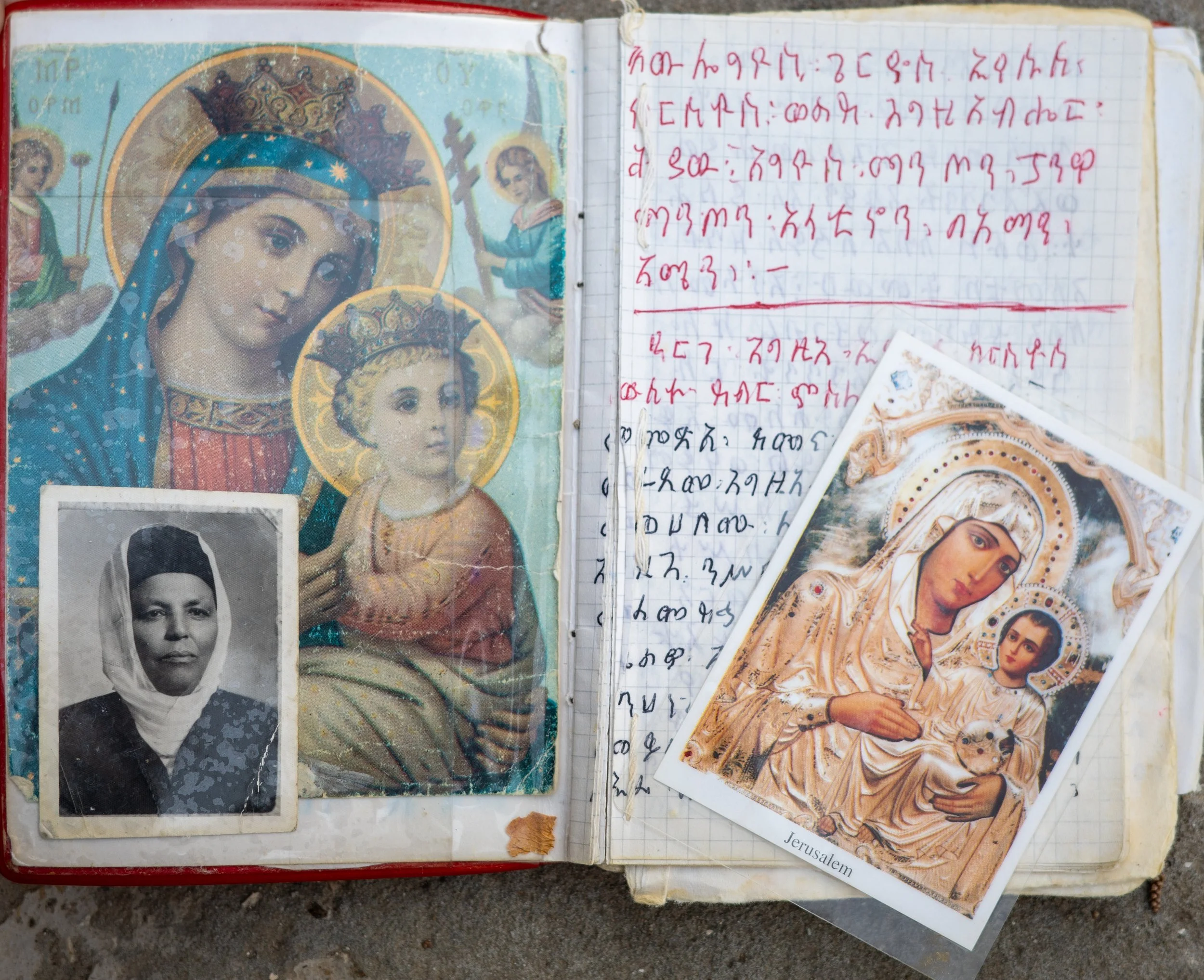 Open notebook with religious images, including icons of Mary and baby Jesus, a card with the word Jerusalem, and a black-and-white photo of a woman, alongside handwritten text in a language with characters resembling Ge'ez script.