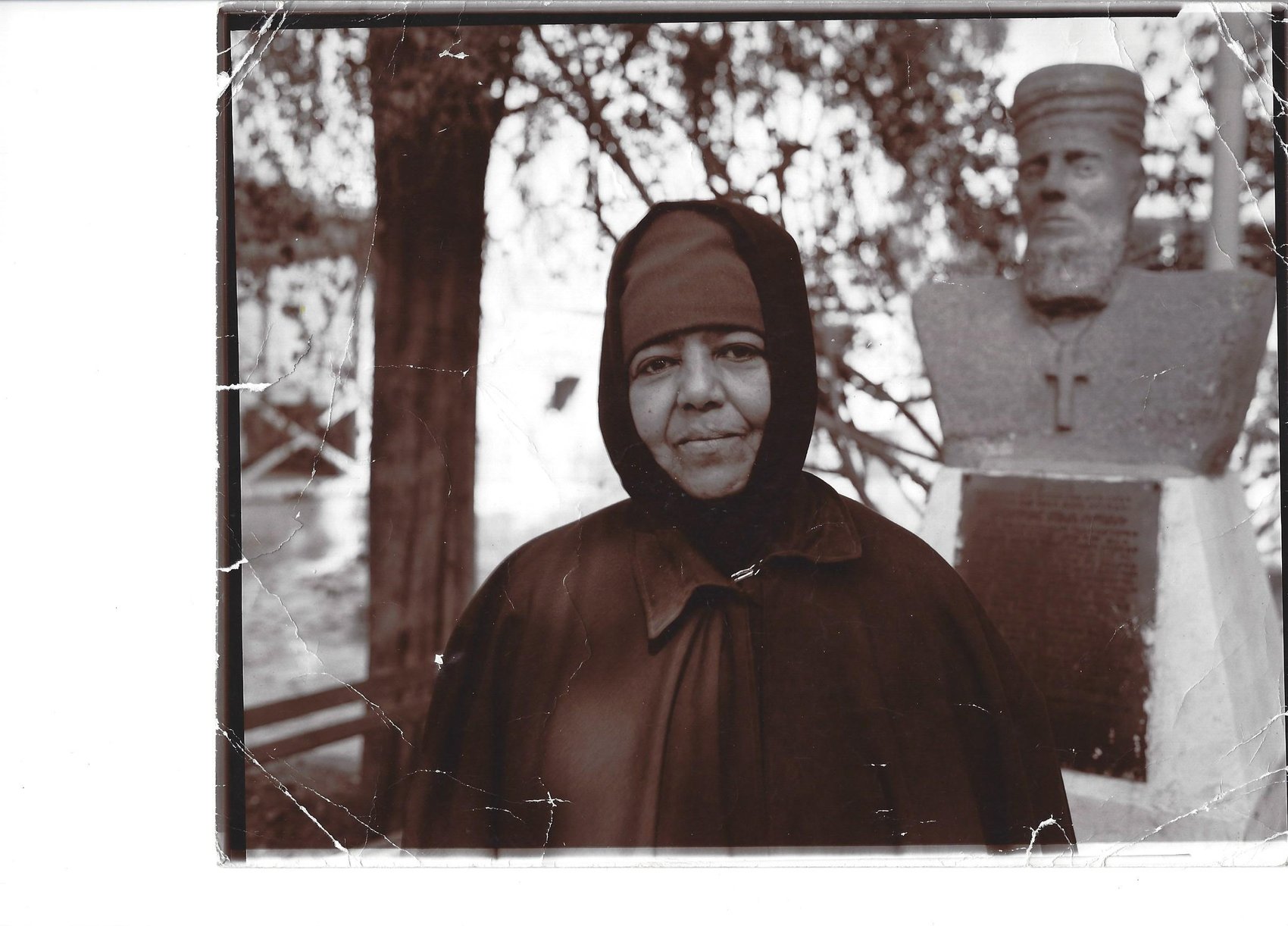 A black and white photograph of a woman wearing a headscarf and coat, standing outdoors near a bust sculpture of a man with a beard and a hat, with a large tree in the background. The photo shows signs of wear and tear.
