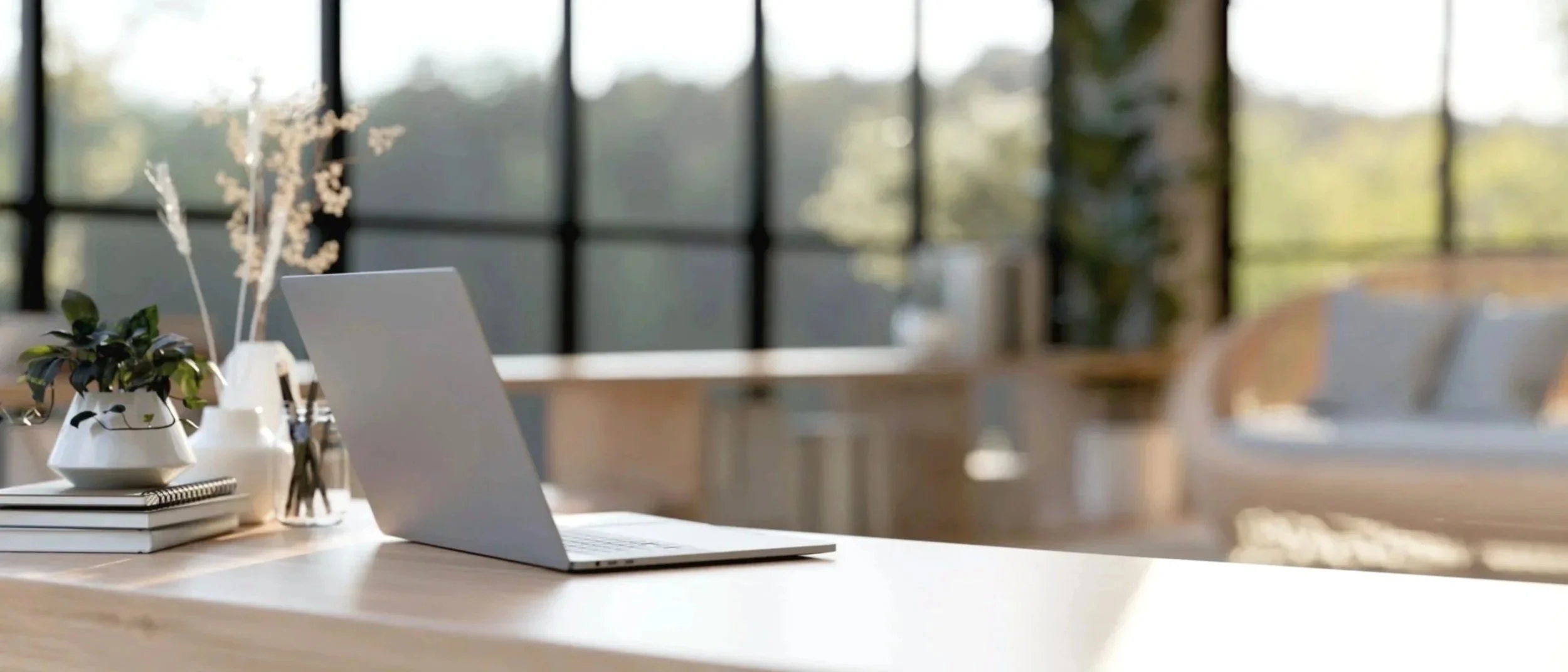 Desk with open laptop, potted plant, notebooks, and decorative items in a bright office with large windows and outdoor view.