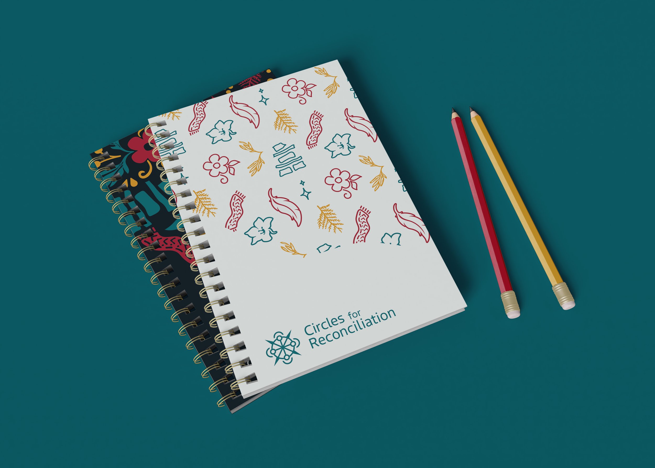 Two spiral notebooks with black and white covers and gold rings, and two pencils, one red and one yellow, on a teal surface. The white notebook has colorful floral and leaf illustrations with the text 'Circles for Reconciliation' and a logo.