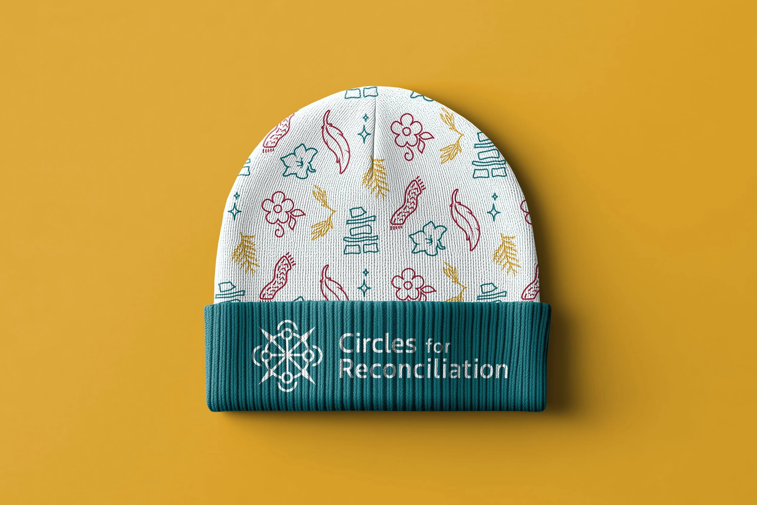 White knit beanie with colorful floral and leaf patterns and a teal cuff that says 'Circles for Reconciliation' against a mustard yellow background.