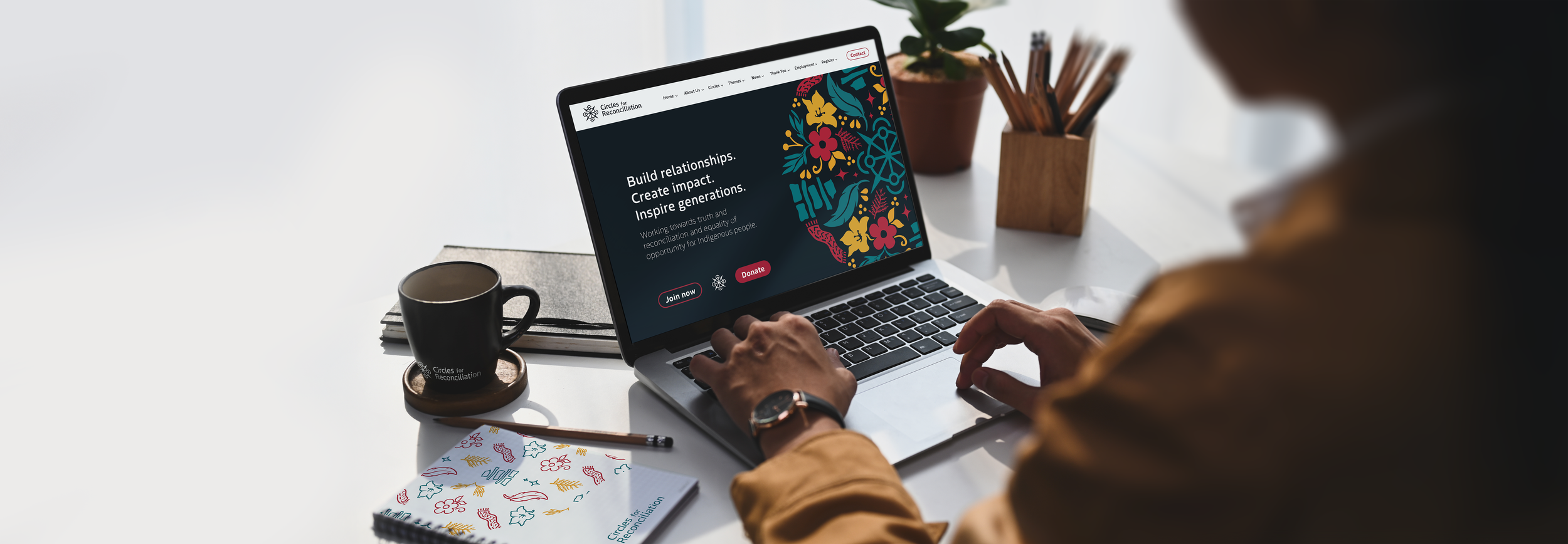 Person working on a laptop at a white desk with a black coffee mug, a notebook, and a potted plant. The laptop screen displays a website titled 'Circles for Reconciliation' with options to join, donate, and learn more.