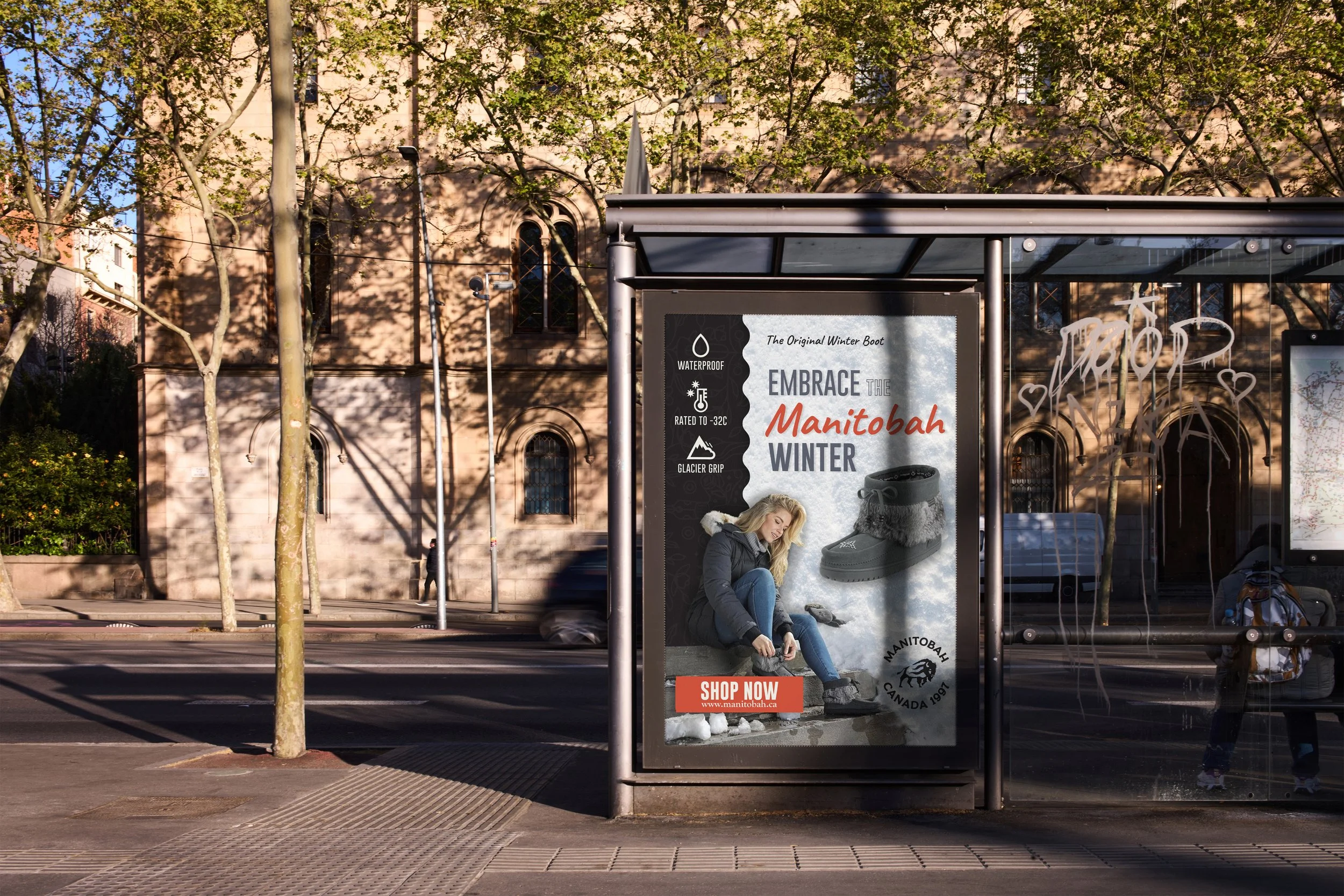 Bus stop advertisement for Manitobah winter footwear, featuring a woman sitting in snow and a pair of black winter boots with fur trim, with text promoting waterproof, cold-rated, glacier grip boots, and a 'Shop Now' button.