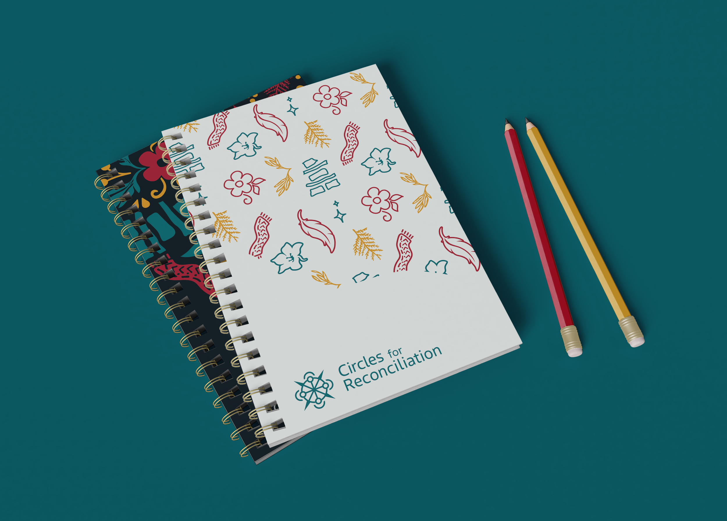 Two spiral-bound notebooks with floral and abstract designs on the covers, placed on a teal surface, accompanied by a red pencil and a yellow pencil.