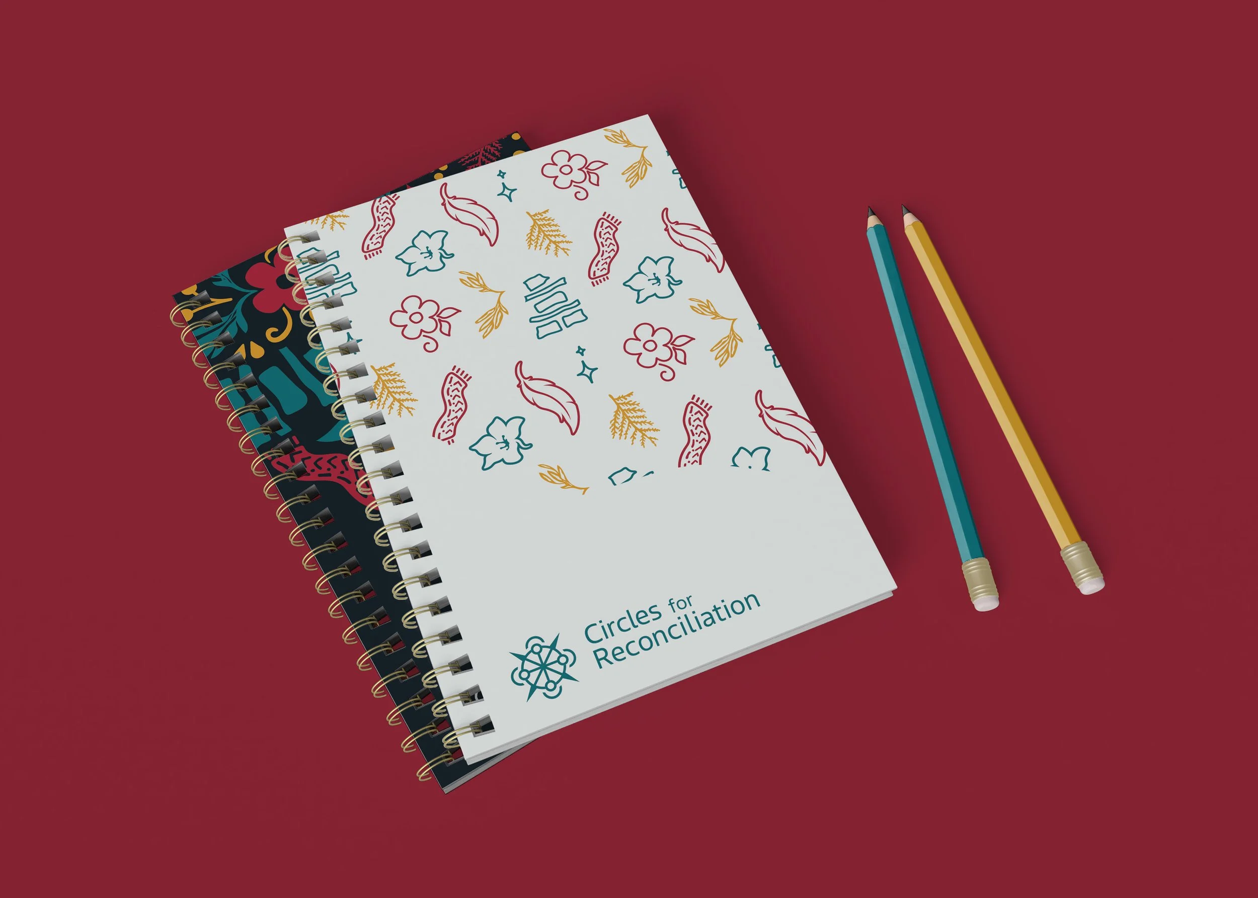 A set of three notebooks with decorative covers and the logo 'Circles for Reconciliation' on the top notebook, along with two colored pencils on a red surface.