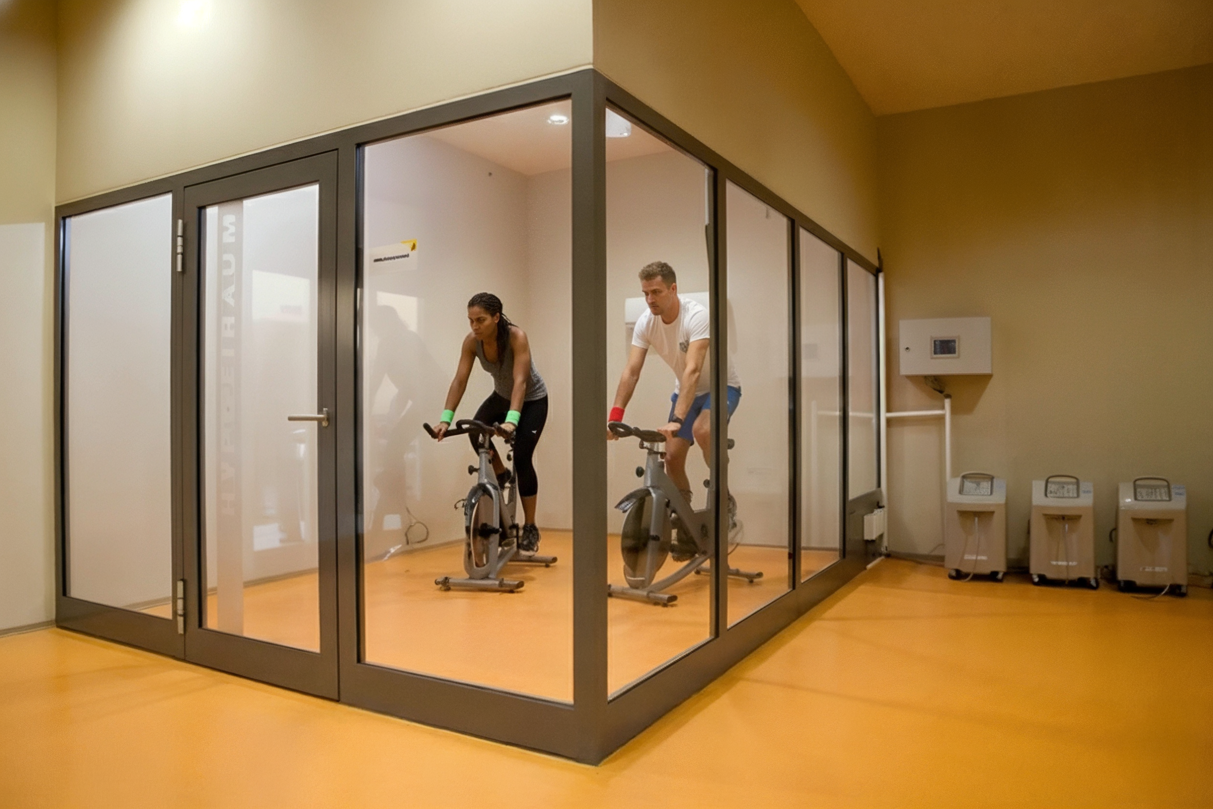 Zwei Menschen trainieren auf stationären Fahrrädern in einem separaten Raum im Fitnessstudio.