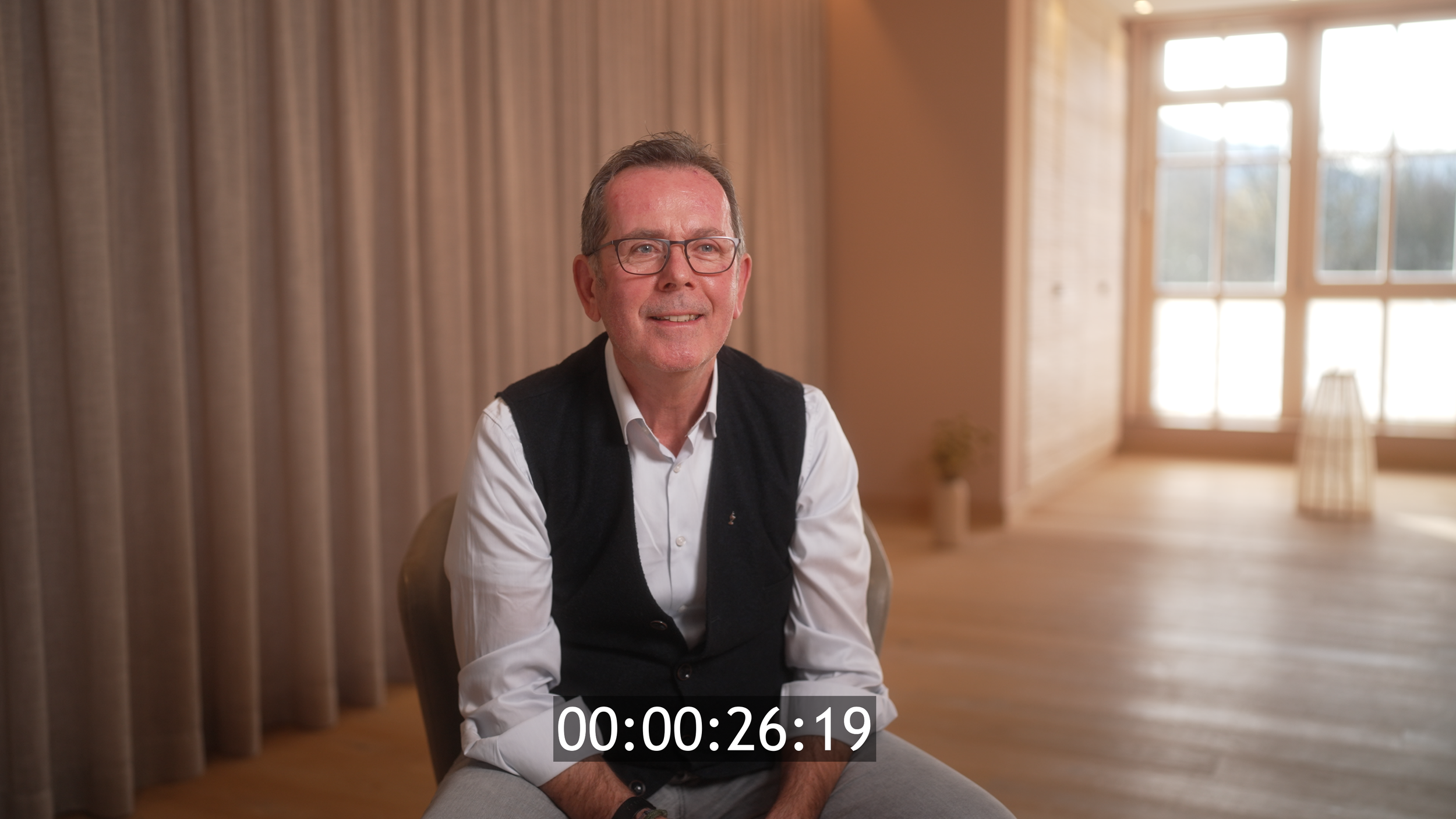 Ein Mann sitzt in einem hellen, holzverkleideten Raum vor einem großen Fenster und lächelt. Er trägt eine weiße Hemd und eine schwarze Weste, trägt Brille und hat kurze Haare.