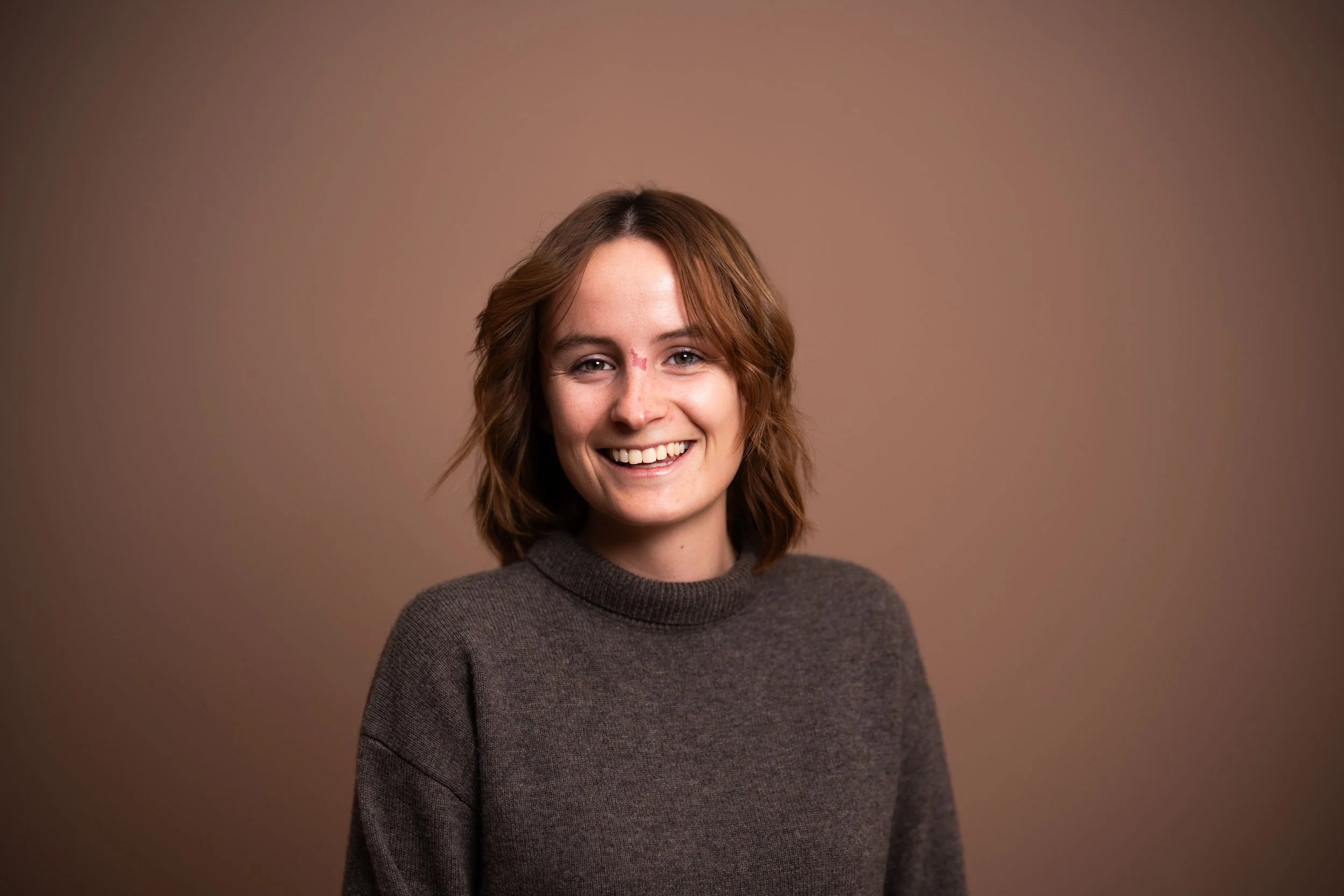 Lächelnde junge Frau mit braunen, gewellten Haaren, trägt einen grauen Pullover, vor neutralem braunem Hintergrund.