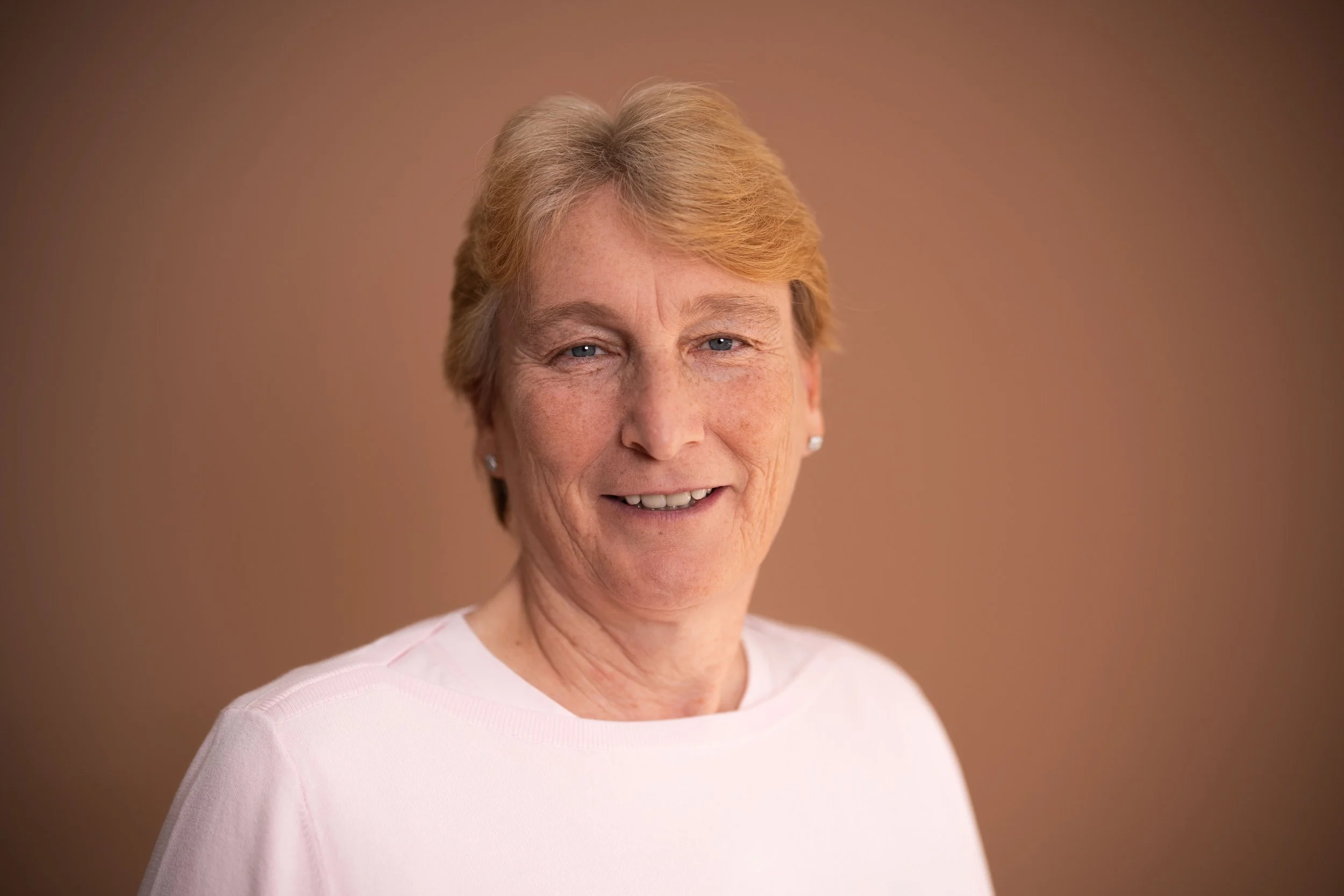 Porträt einer älteren Frau mit kurzen, blonden Haaren, blauer Augen, trägt weiße Kleidung und kleine Ohrringe, vor einem braunen Hintergrund.