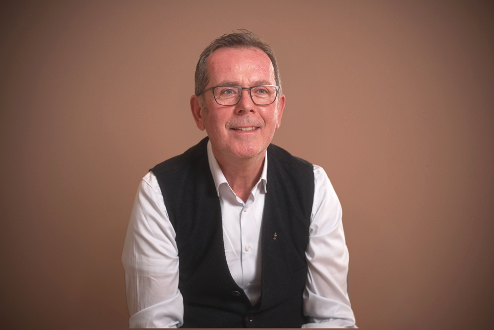 Ein älterer Mann mit Brille, im weißen Hemd und schwarzer Weste, lächelnd, vor einem einfarbigen Hintergrund.