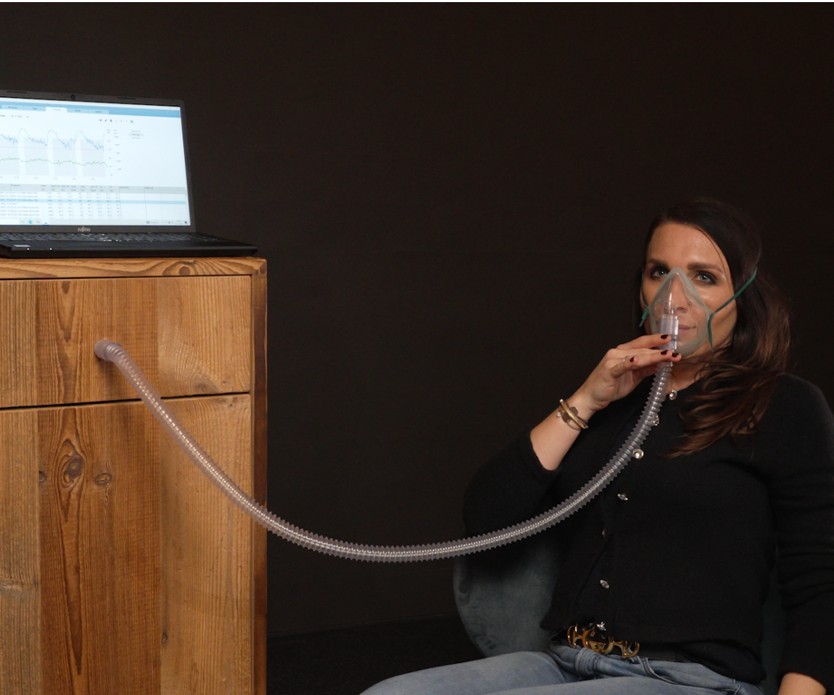 Frau sitzt mit einer Atemmaske und verbundenem Schlauch neben einem Laptop, der auf einem Holzschrank steht, und überwacht eine Datenanalyse auf dem Bildschirm.