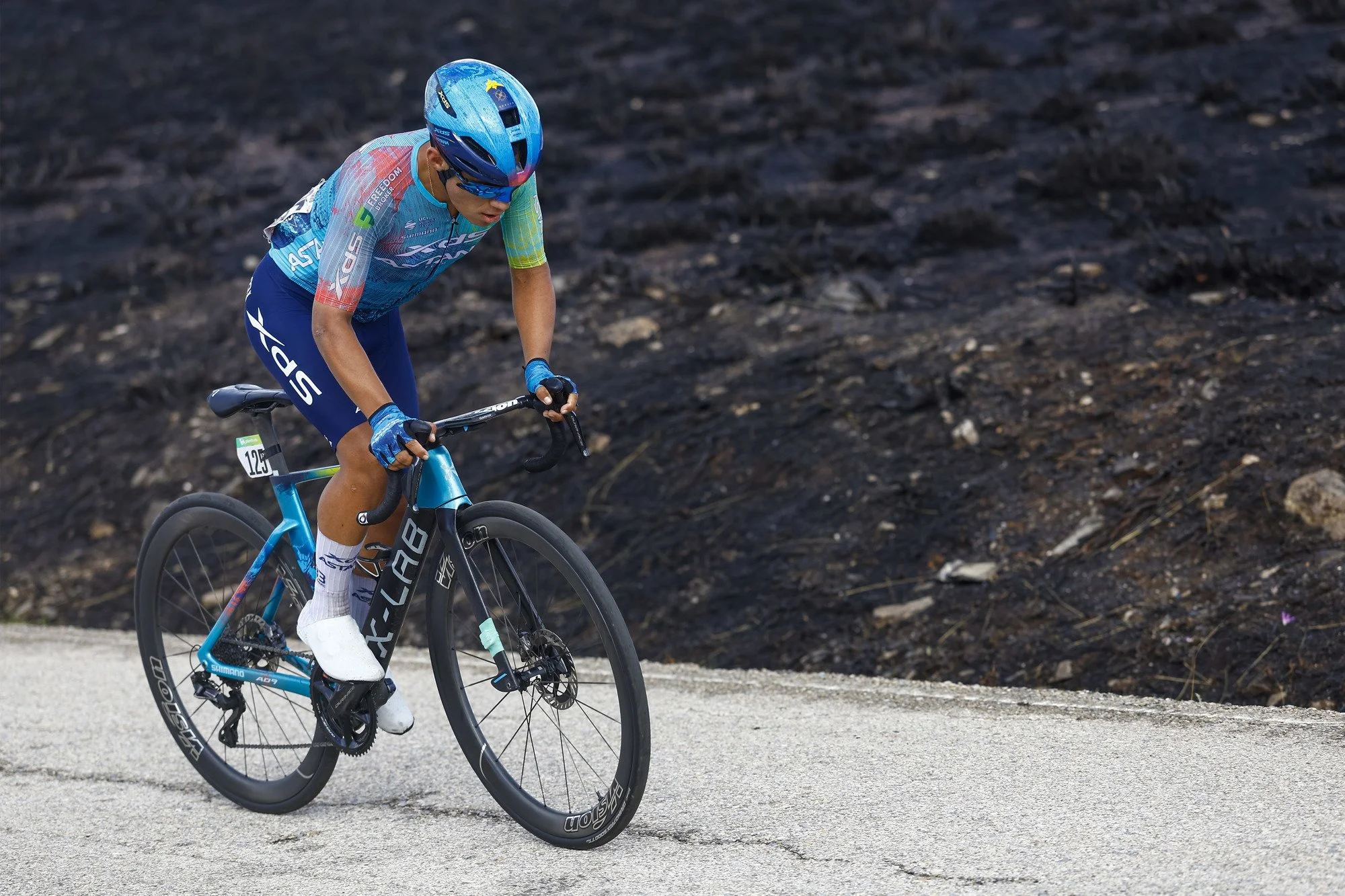 Radfahrer in blauer Kleidung und Helm fährt auf einem Asphaltweg, im Hintergrund schwarzer Boden.
