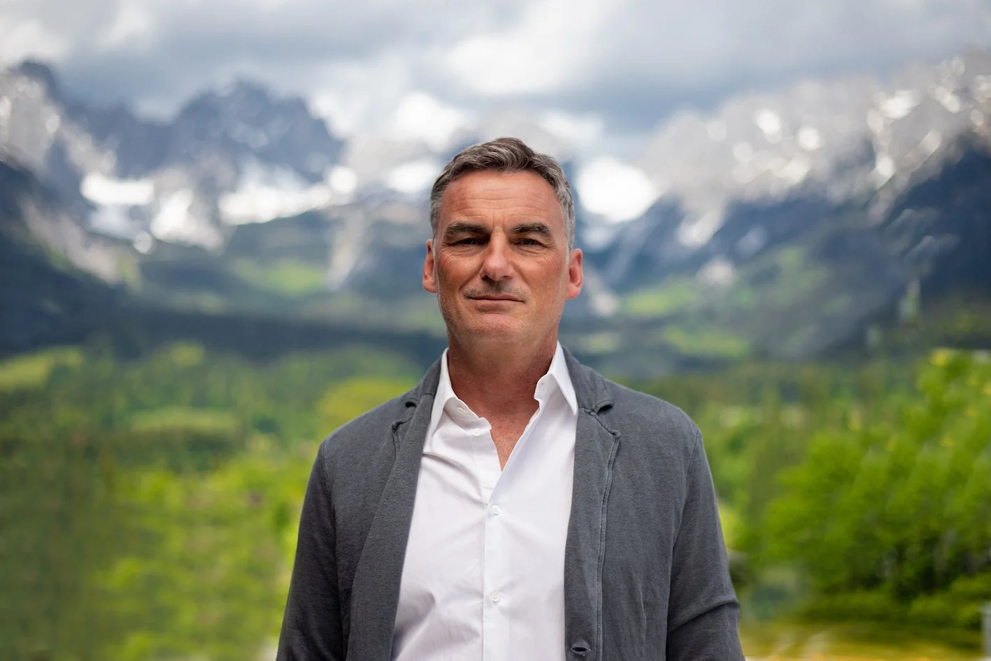 Ein Mann mit grauem Haar trägt einen weißen Hemd und eine graue Jacke vor einer Berglandschaft mit schneebedeckten Gipfeln und grüner Natur im Hintergrund.