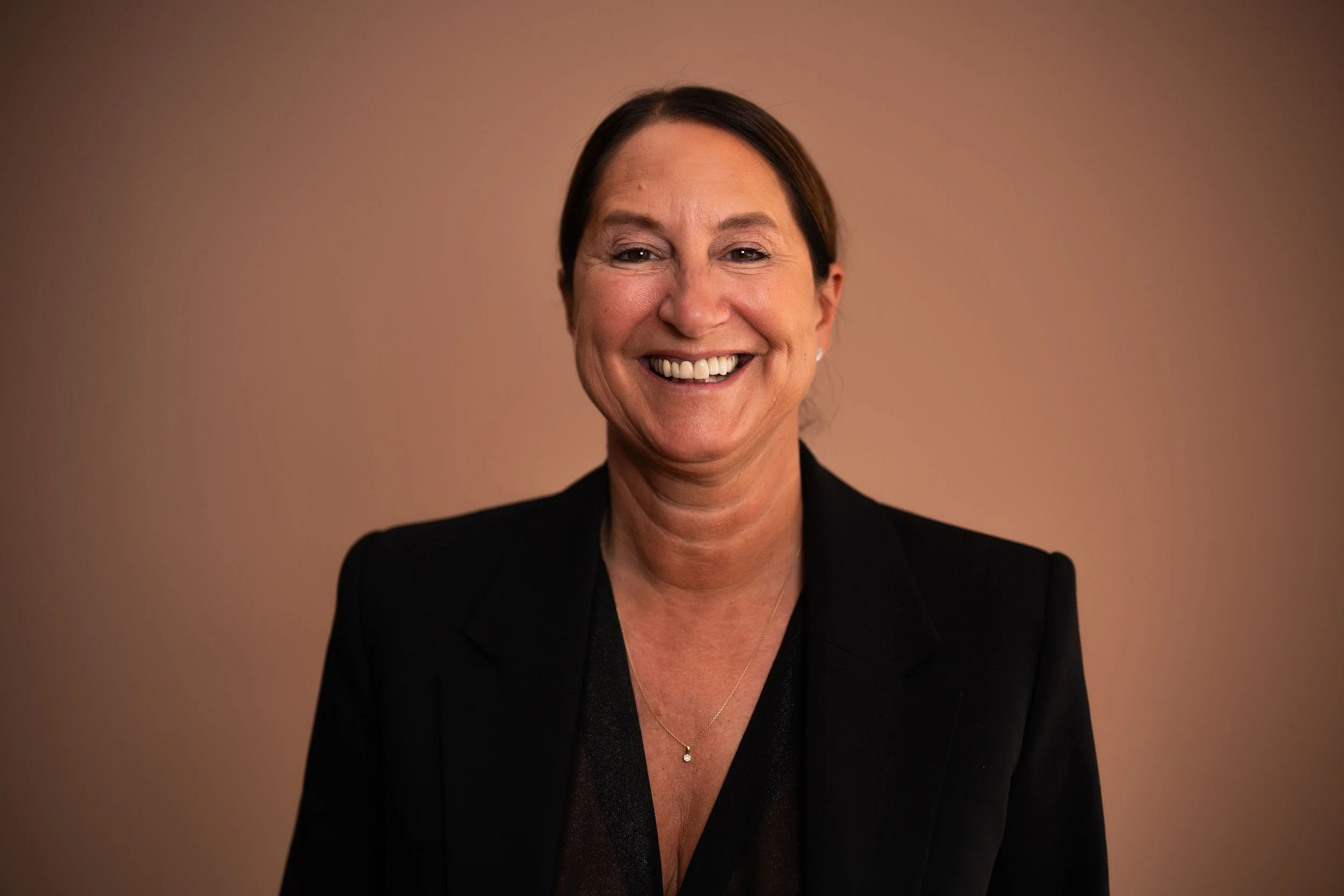 Porträt einer lächelnden Frau in dunklem Blazer vor einem unscharfen braunen Hintergrund.