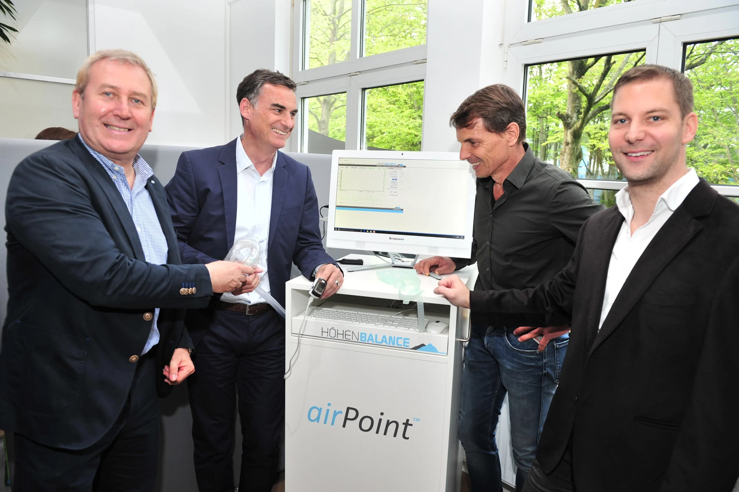 Vier Männer in Anzügen, die an einem medizinischen Gerät mit einem Monitor arbeiten, in einem hellen Raum mit großen Fenstern und grünen Bäumen im Hintergrund.