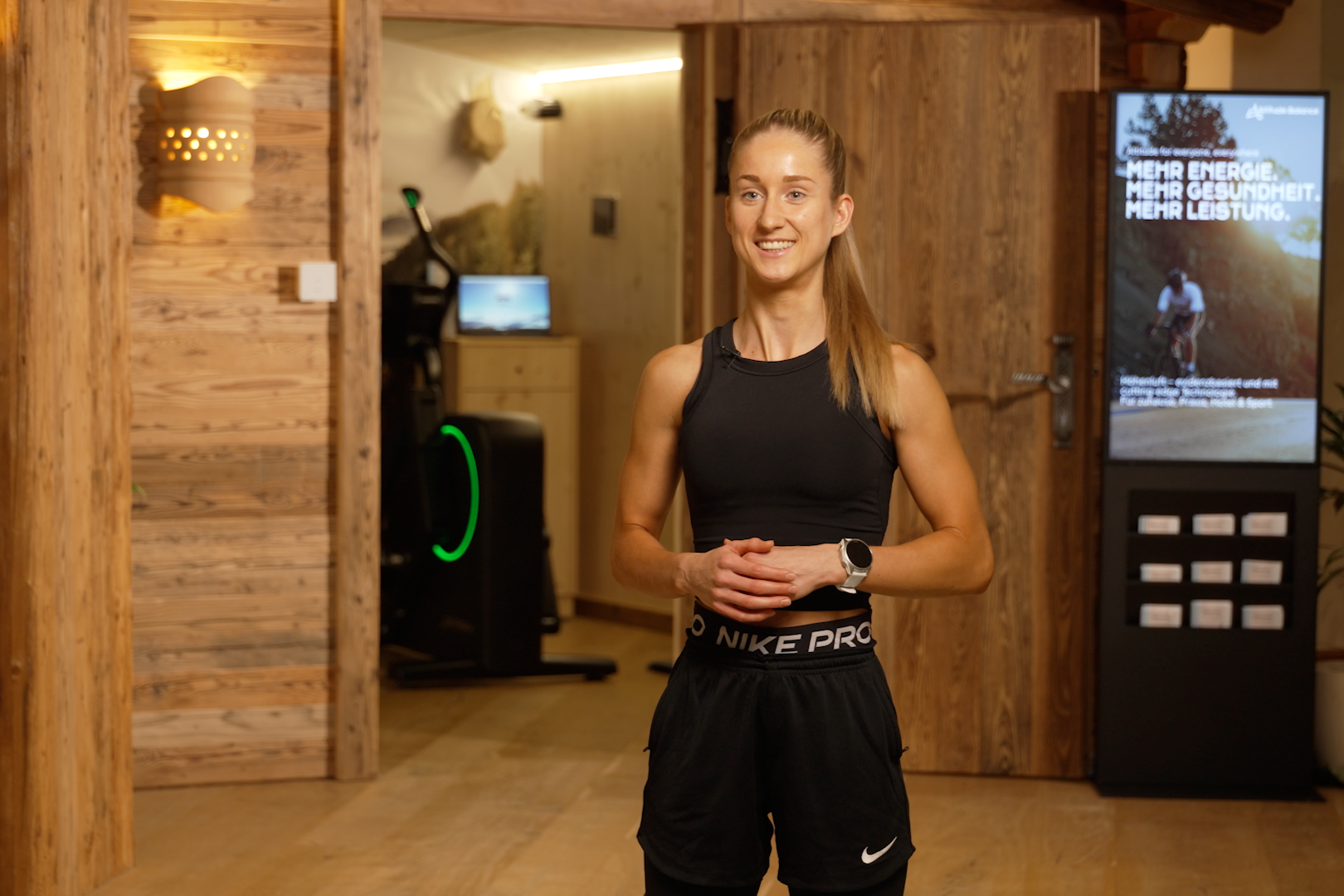 Eine lächelnde jüngere Frau in sportlicher Kleidung, darunter ein schwarzes Top und eine Nike Pro Shorts, steht in einem Holzraum mit moderner Einrichtung, vermutlich in einem Fitnessstudio oder Wellnessbereich.
