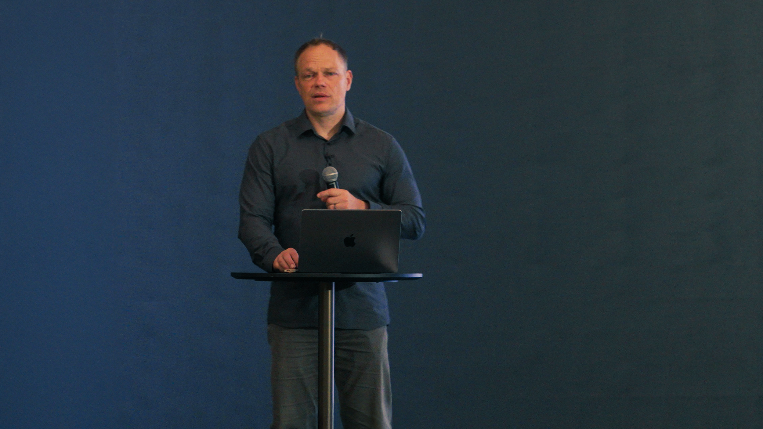 Mann hält Mikrofon vor einem Laptop auf einem Stehtisch, befindet sich in einem Raum mit dunklem Hintergrund.