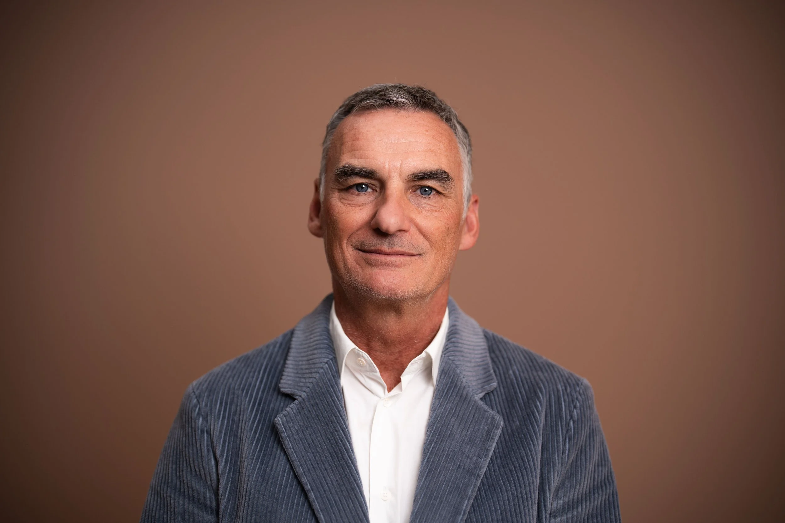 Nahaufnahme eines lächelnden Mannes mit grauem Haar, weißem Hemd und grauem Jackett vor braunem Hintergrund.