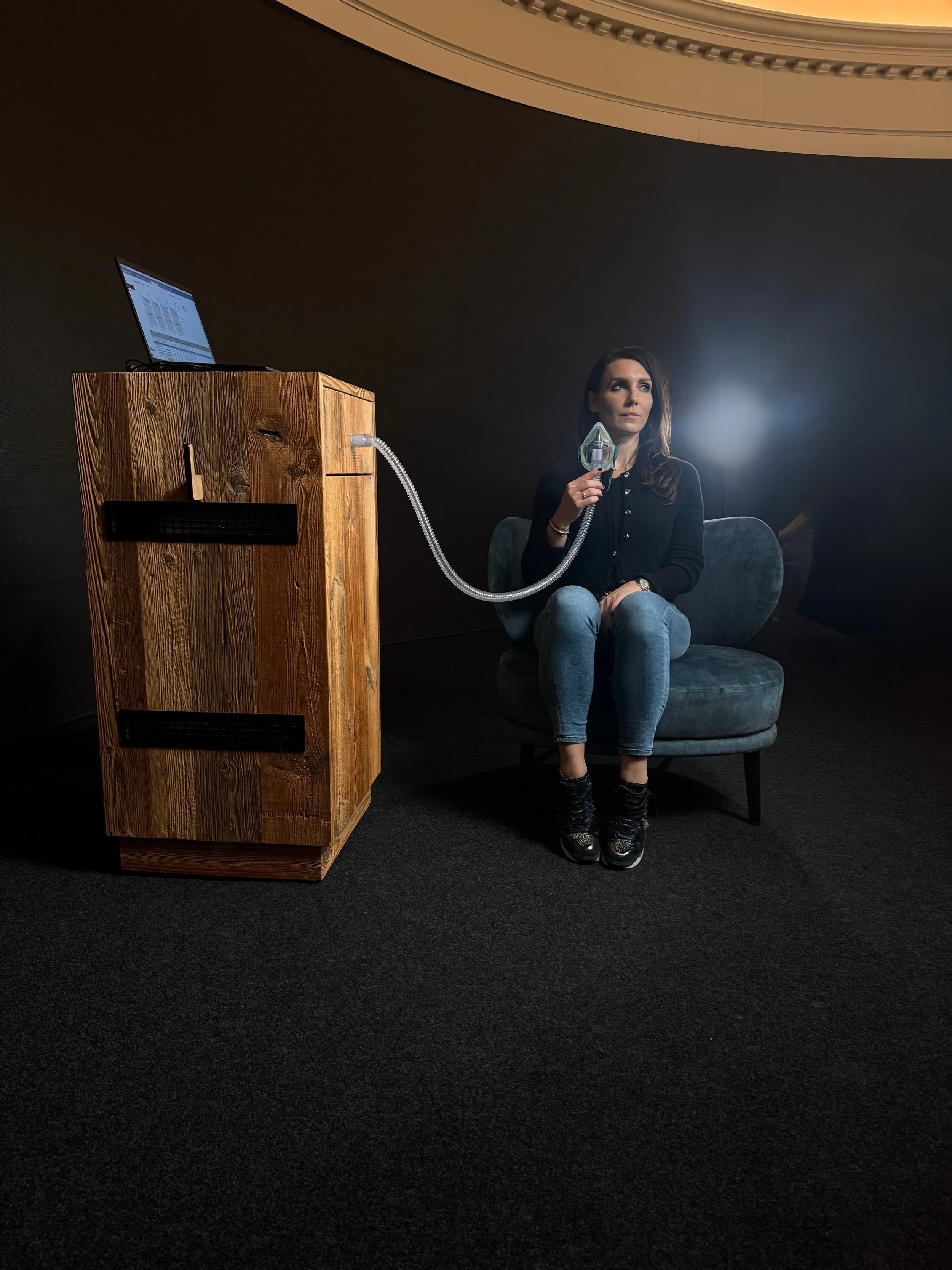 Frau sitzt auf einem dunklen Sessel, hält eine Atemmaske an den Mund, dabei sind sowohl der Schlauch als auch ein Laptop auf einem Holzpult zu sehen.