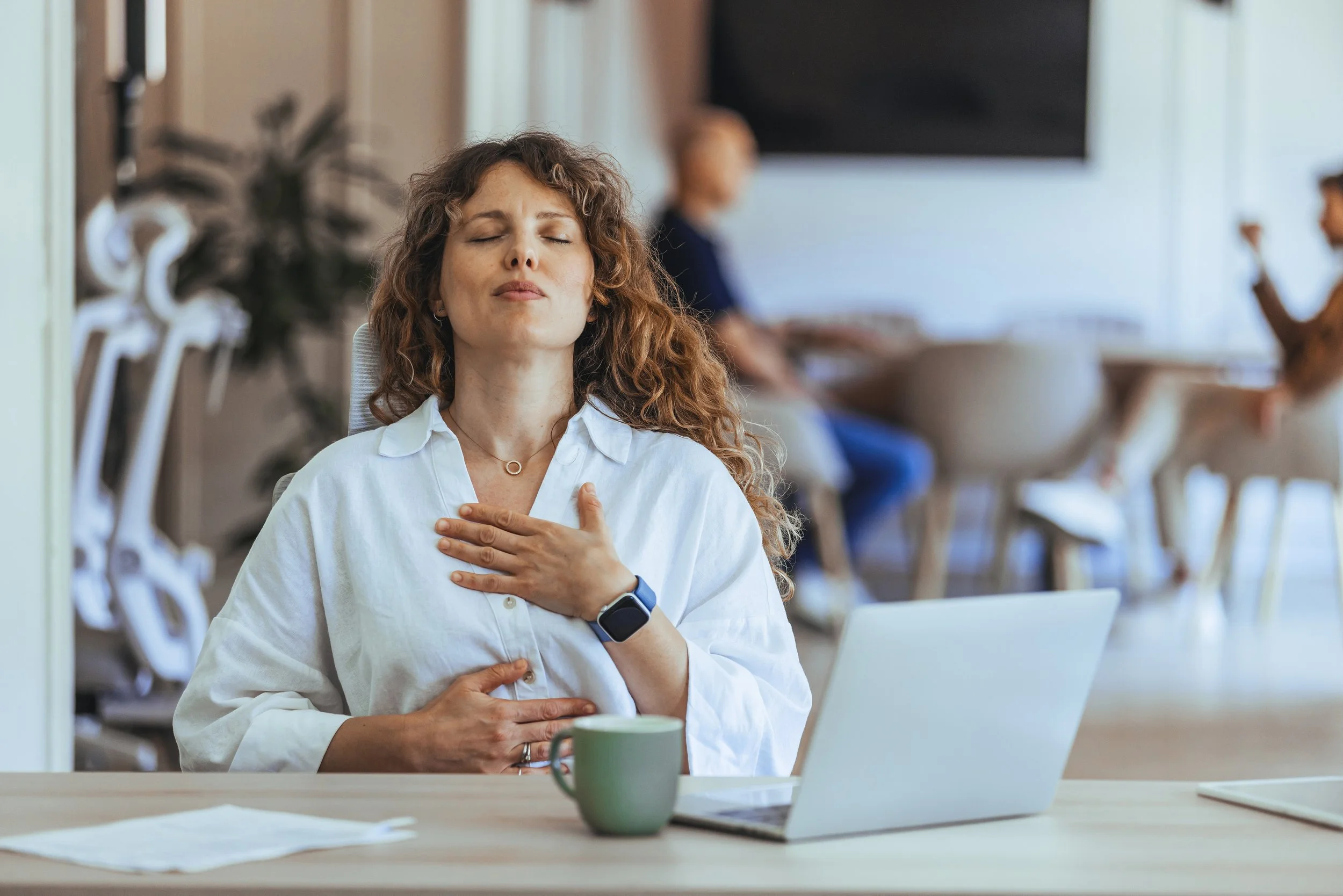 Frau in einem weißen Hemd sitzt an einem Tisch mit geschlossenen Augen, die rechte Hand auf der Brust, die linke Hand auf dem Bauch, neben einem Laptop und einer grünen Tasse, im Hintergrund sitzen Menschen in einem modernen Raum.