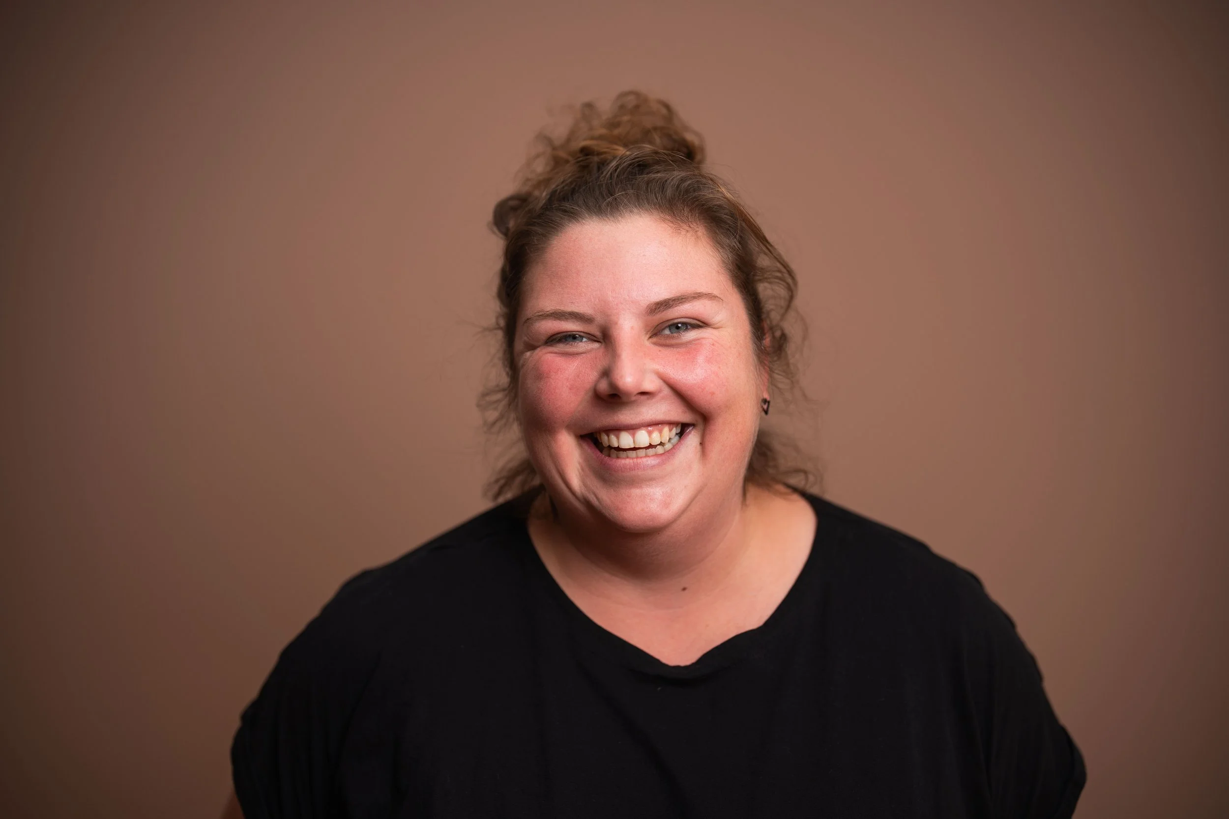 Lächelnde Frau mit braunen, lockigen Haaren vor einem braunen Hintergrund.