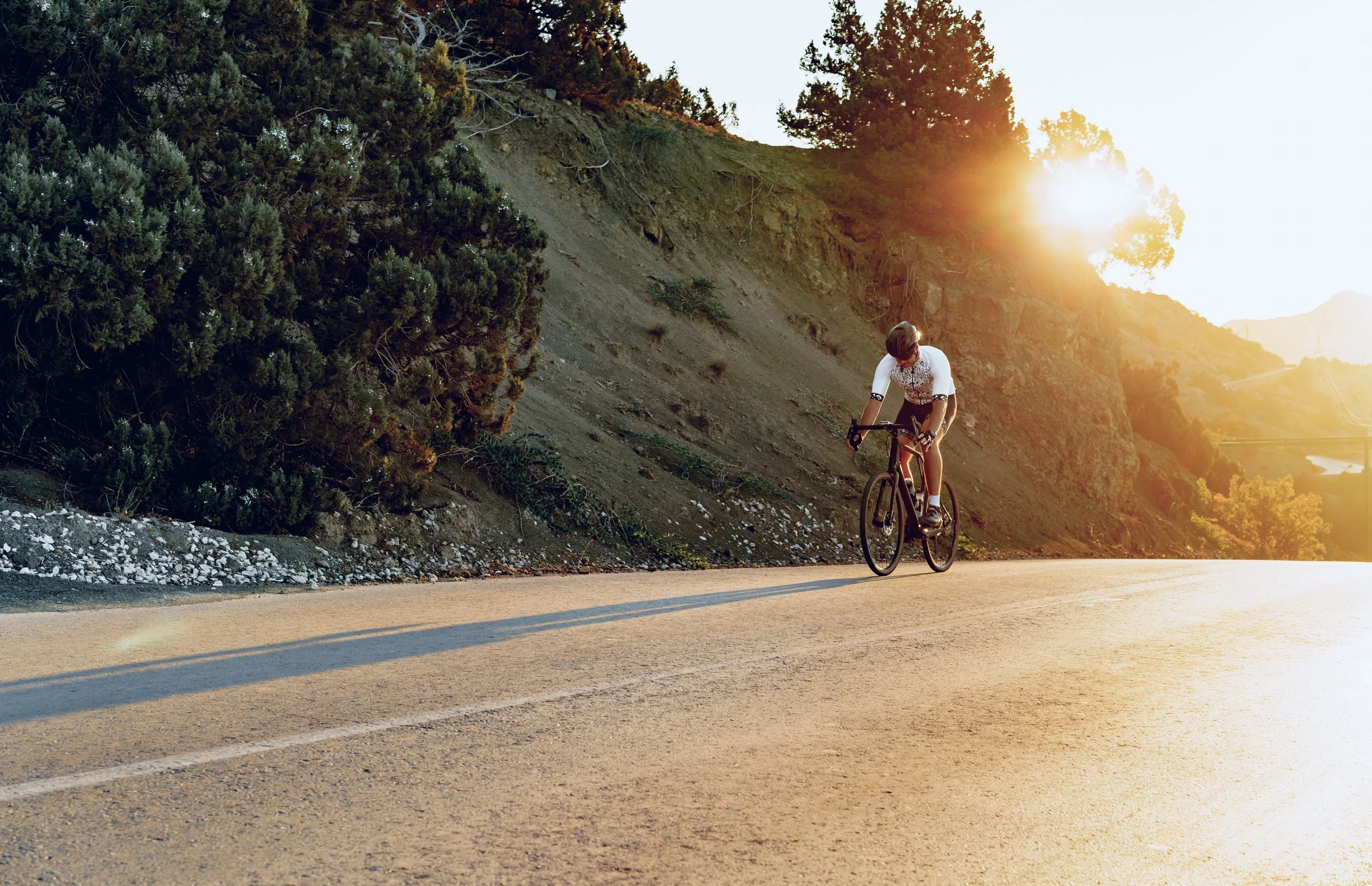 Ein Radfahrer fährt auf einer Landstraße im Sonnenuntergang, umgeben von Hügeln und Bäumen.