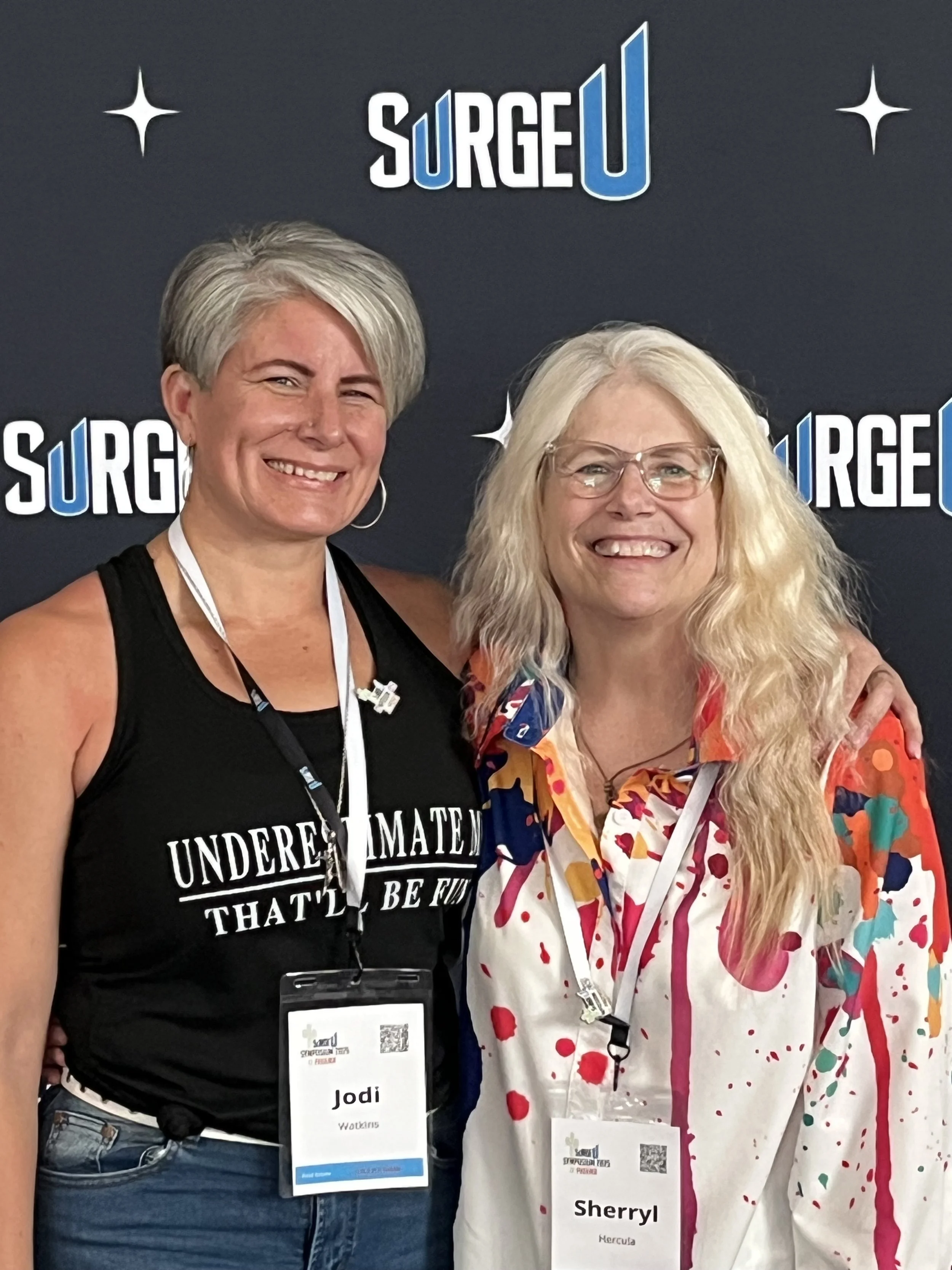 Two women smiling, standing close together in front of a backdrop with the logo 'SurgeU.' The woman on the left has short, gray hair, a black tank top with whitespace text, and a name tag labeled 'Jodi Watkins.' The woman on the right has long, curly blonde hair, glasses, a colorful, splattered pattern shirt, and a name tag labeled 'Sherryl Mercía.'