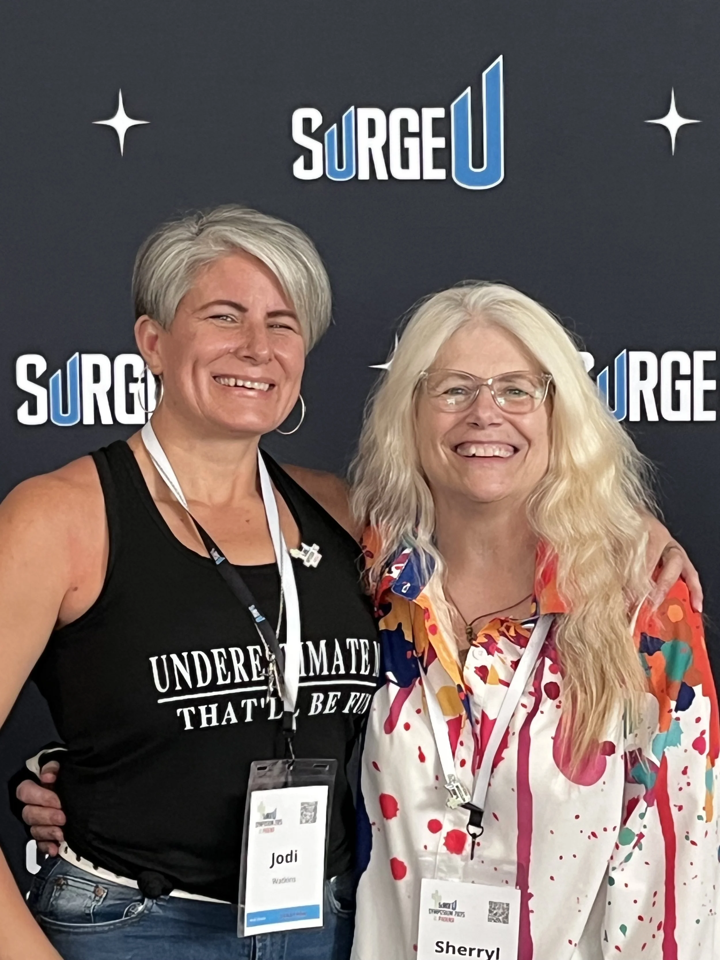 Two smiling women standing together at a conference, wearing name tags, in front of a backdrop with the logo 'SURGE U' repeated in the background.