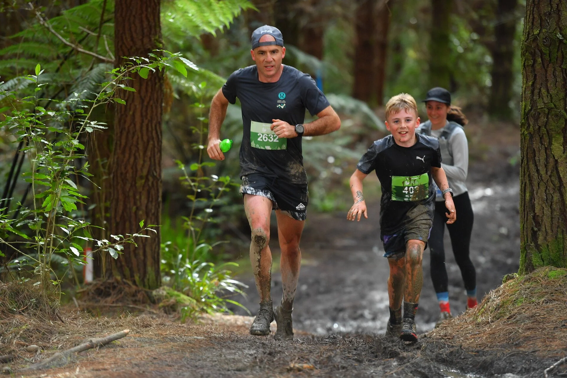 Three people running through a muddy forest trail, including a man and two children, all wearing athletic gear and race bibs.