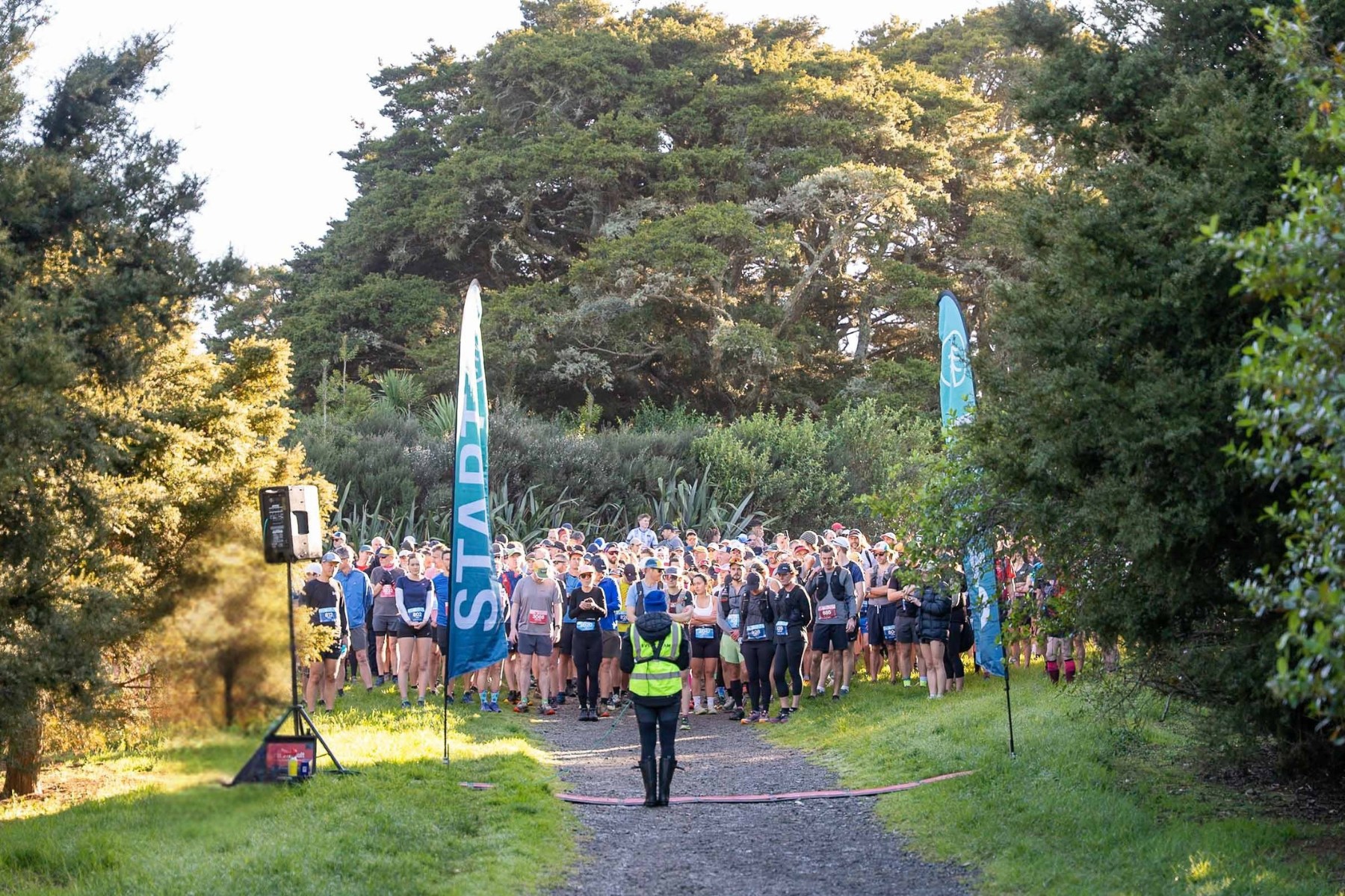 Group of people gathered at the starting line for a race, with race flags and a person in a reflective vest overseeing the event, surrounded by trees and nature.