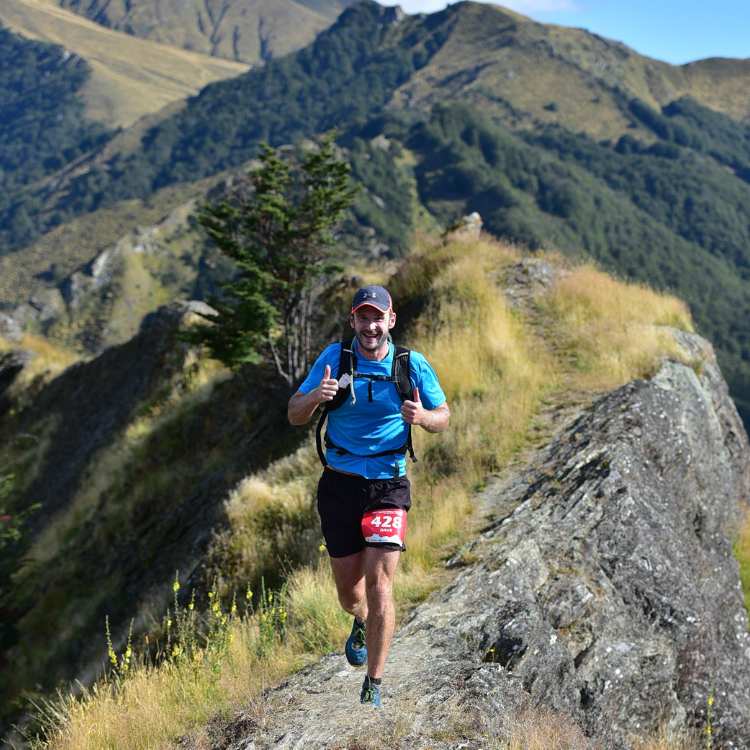 Man running on a mountain trail with a smile, surrounded by mountainous landscape and blue sky.