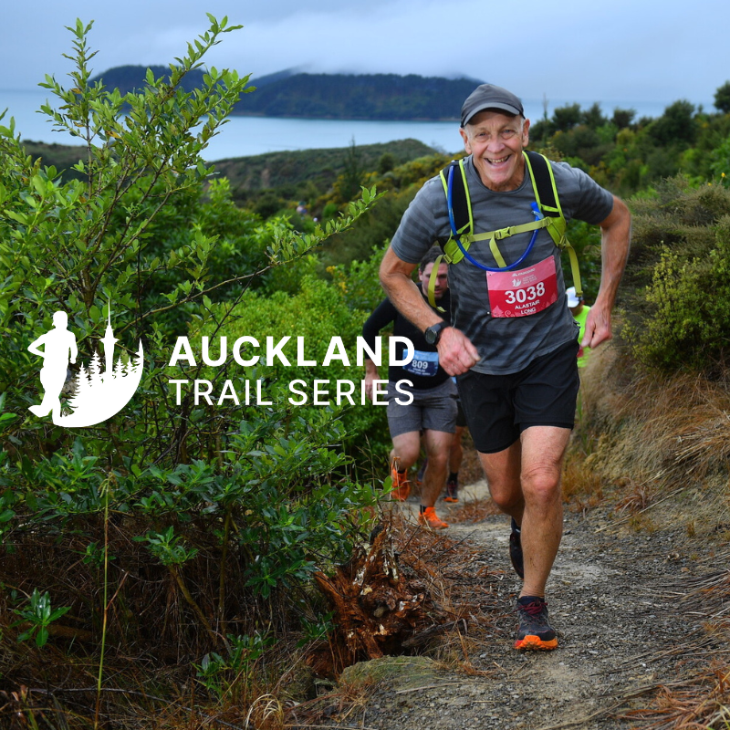 Older man trail running on a dirt path in a scenic outdoor setting with green bushes, a body of water, and mountains in the background, participating in the Auckland Trail Series.