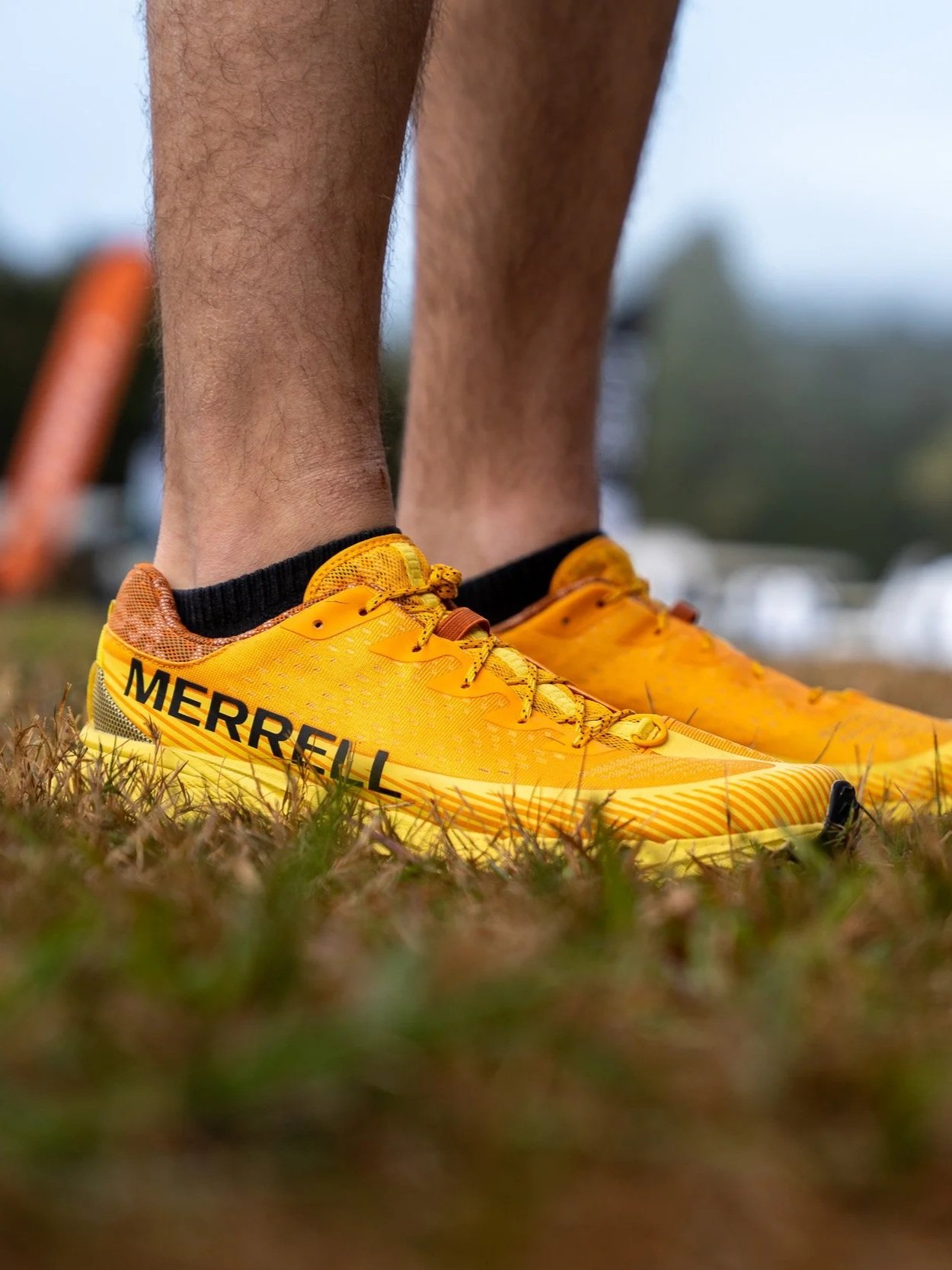 Close-up of yellow Merrell trail running shoes on grass, with a person's lower legs visible, in an outdoor setting.